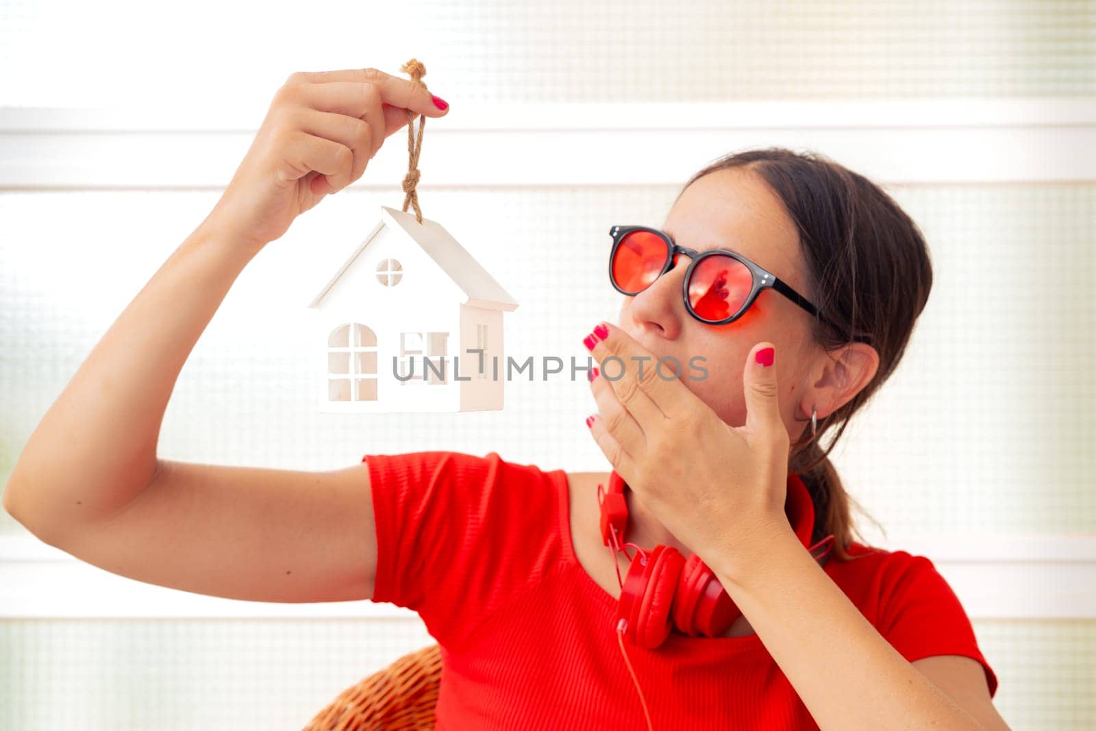 A young woman wearing red sunglasses and headphones holds a small house model while covering her mouth in surprise, displaying an expressive and curious gesture.