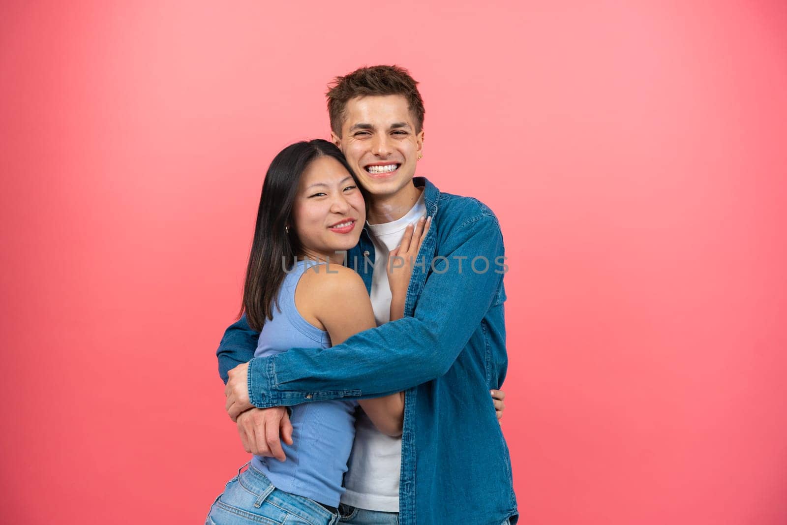 Loving young man hugging his pretty girlfriend on pink studio background, happy multiracial lovers enjoying time together, hugging, looking at camera.