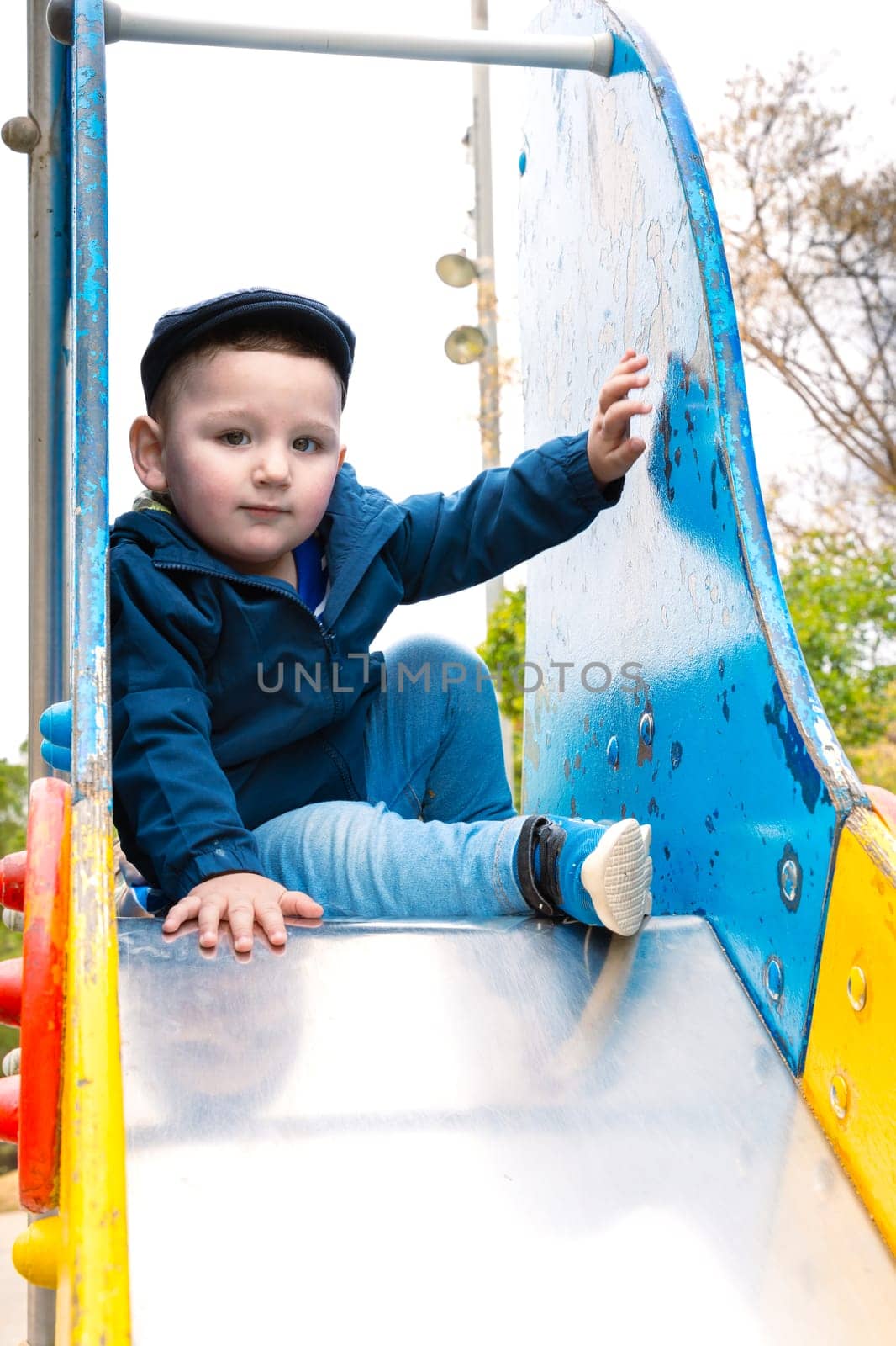 Sweet little Caucasian boy in cap having fun smiling widely sliding down the slide.