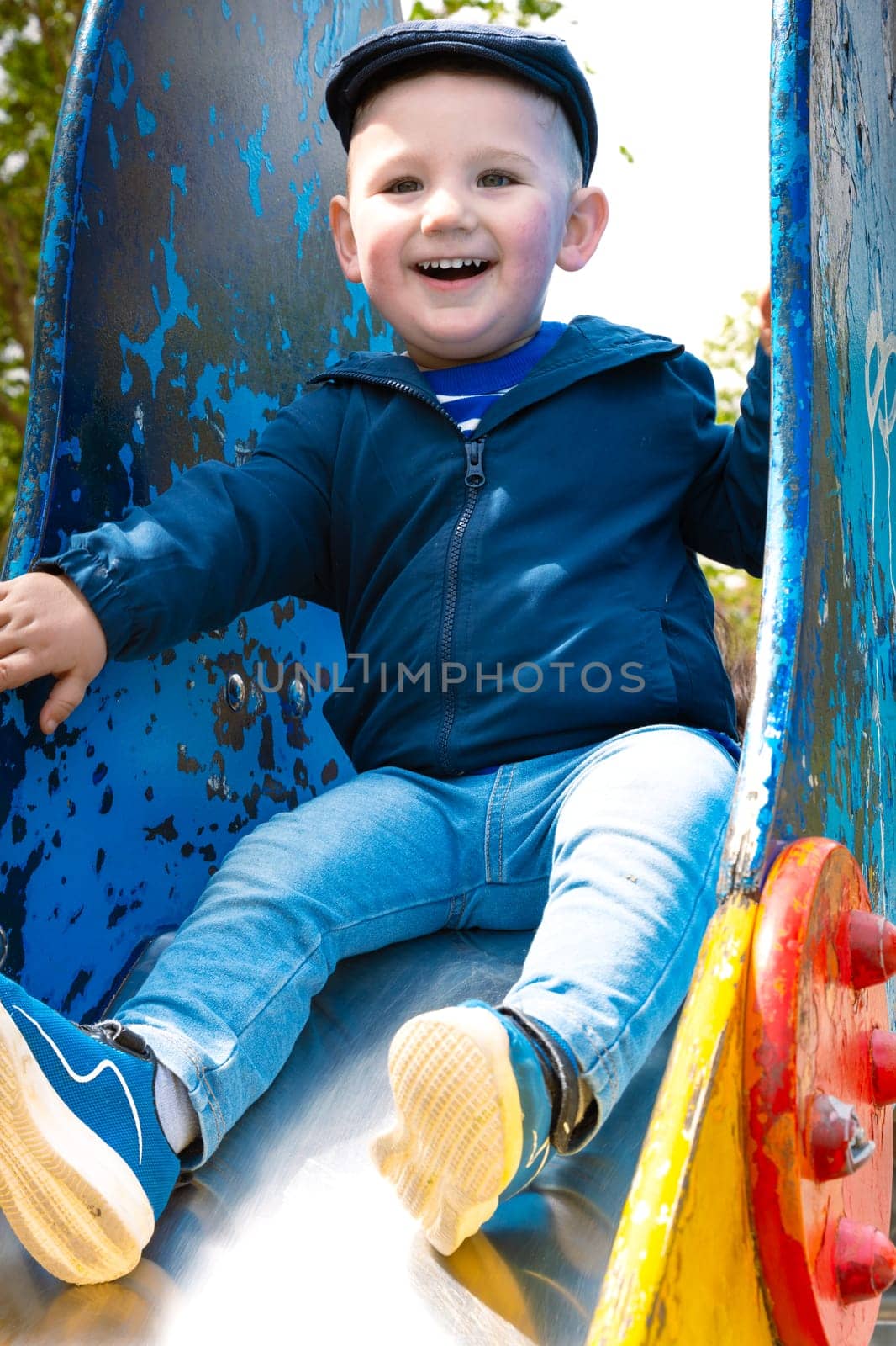 Sweet little Caucasian boy in cap having fun smiling widely sliding down the slide.