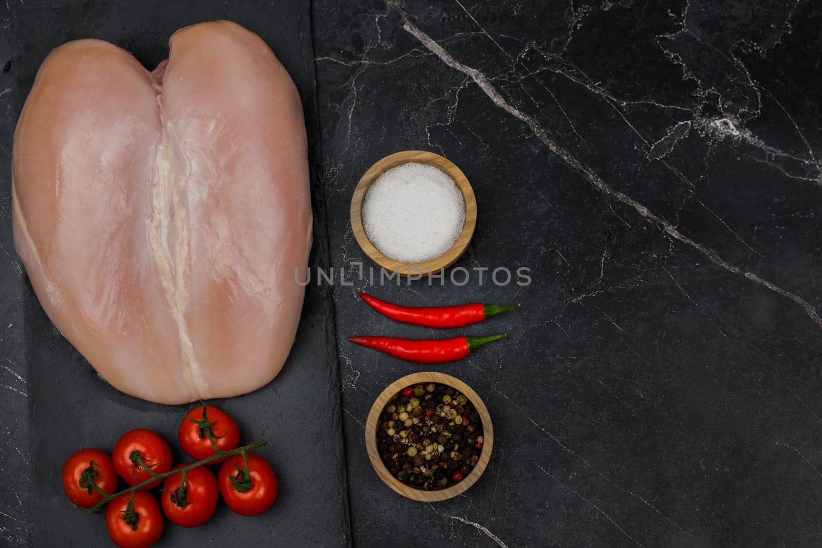 Chicken breast fillet lies on a black board on a dark background with a sprig of tomatoes next to it, spices and salt in wooden bowls on the side, red hot pepper on a dark background with space for text. The concept of proper and healthy nutrition, dietary dishes and recipes. High quality photo