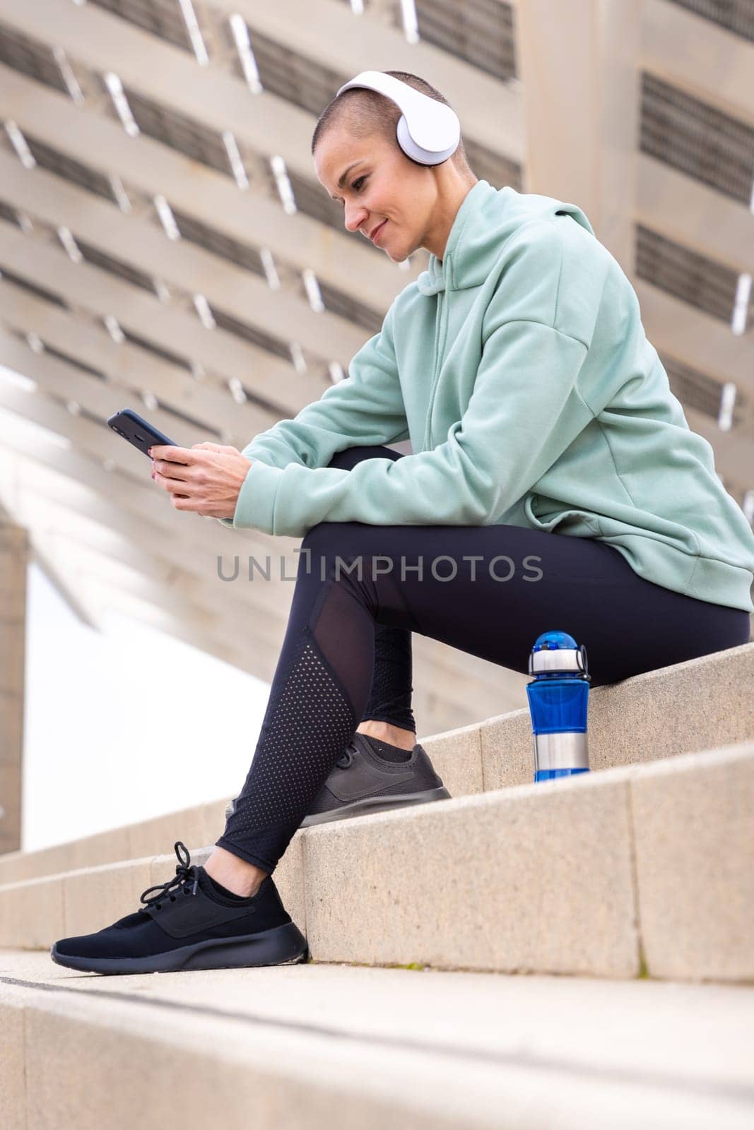 An athletic woman with headphones tired from exercising sits down to take a water break while using a cell phone app.