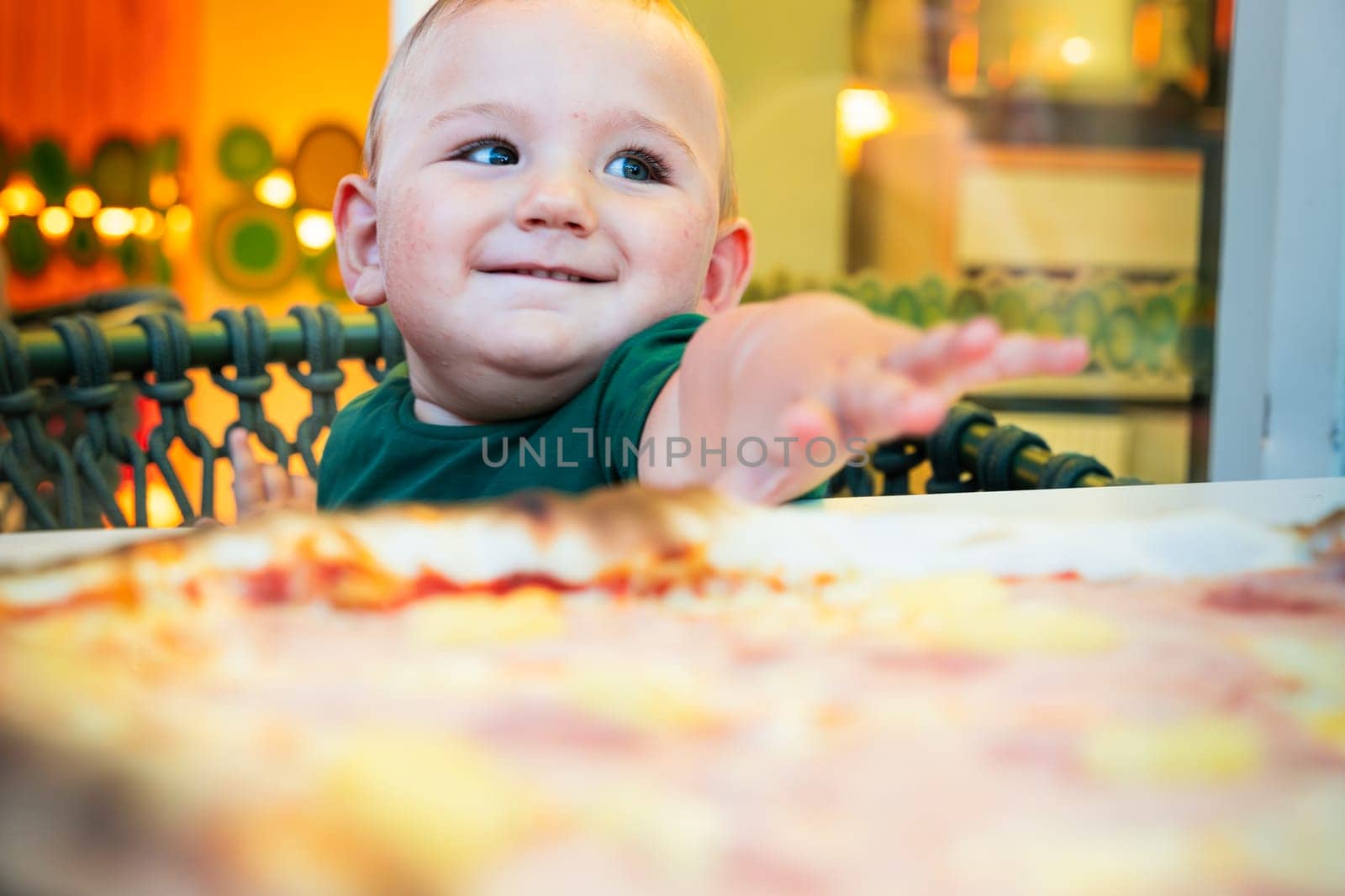 Close-up portrait of a very handsome baby with blonde hair and big green eyes sitting on the terrace of a restaurant, in a chair reaching out to take food from the table.