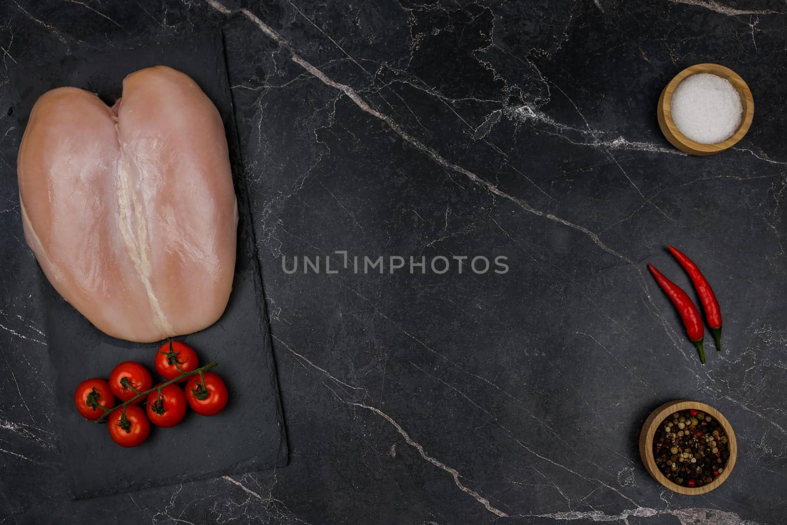 Chicken breast fillet with tomato sprig lies on a black board on the other side with spices and salt in wooden bowls and red hot pepper on a dark background with space for text in the center. The concept of proper and healthy nutrition, dietary dishes and recipes. High quality photo