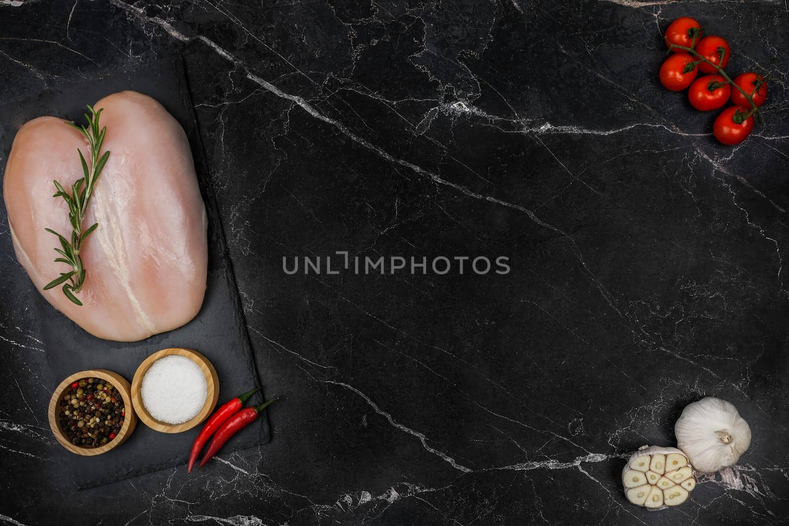 Chicken breast fillet with spices and salt in wooden bowls and red hot pepper lies on a black board on the other side with a sprig of tomato and garlic on a dark background with space for text in the center. The concept of proper and healthy nutrition, dietary dishes and recipes. High quality photo
