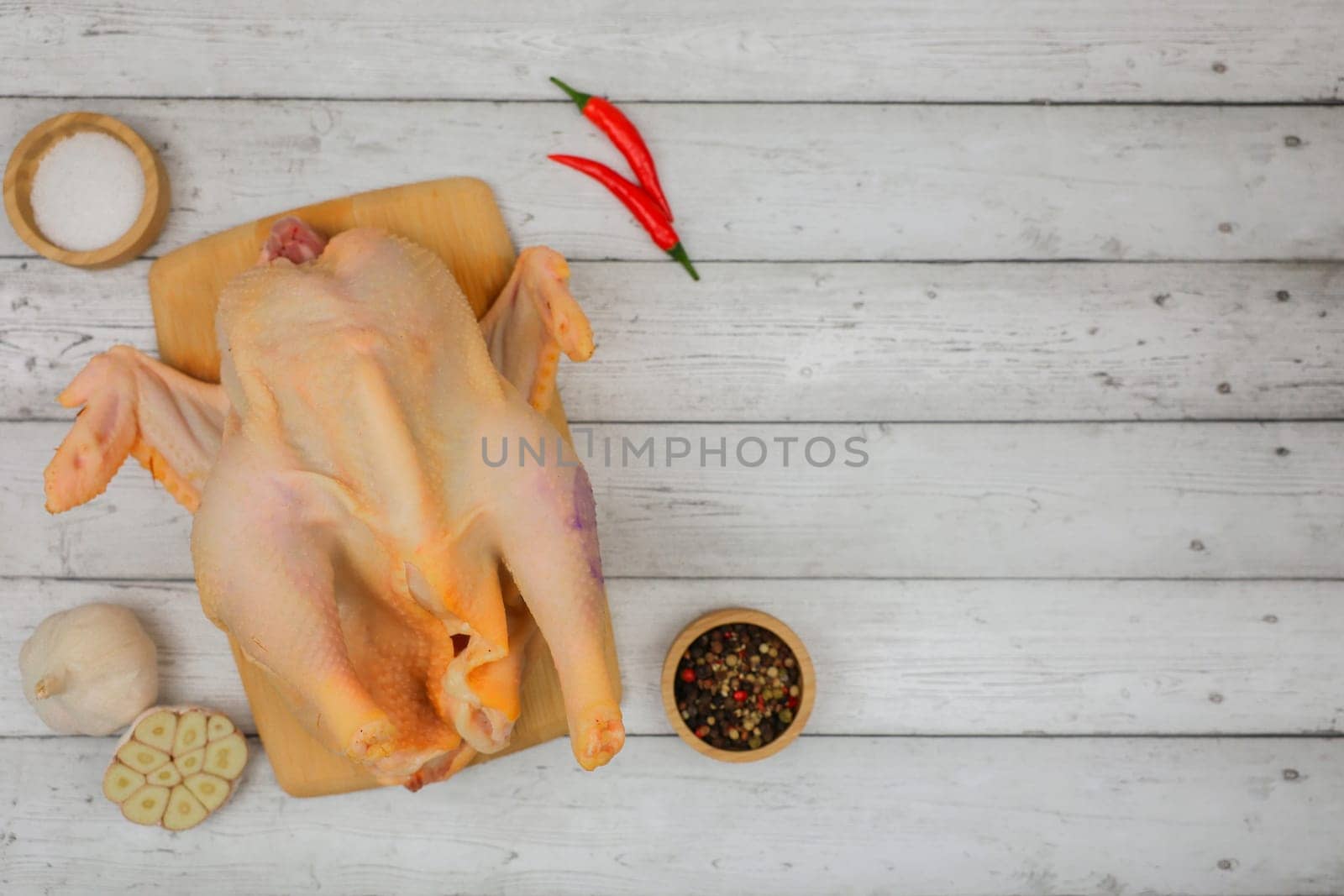 A raw processed chicken carcass lies on a white wooden background on a wooden board with spices and salt in wooden bowls, garlic and red pepper with space for text. The concept of cooking, wholesome and healthy food, recipes. High quality photo