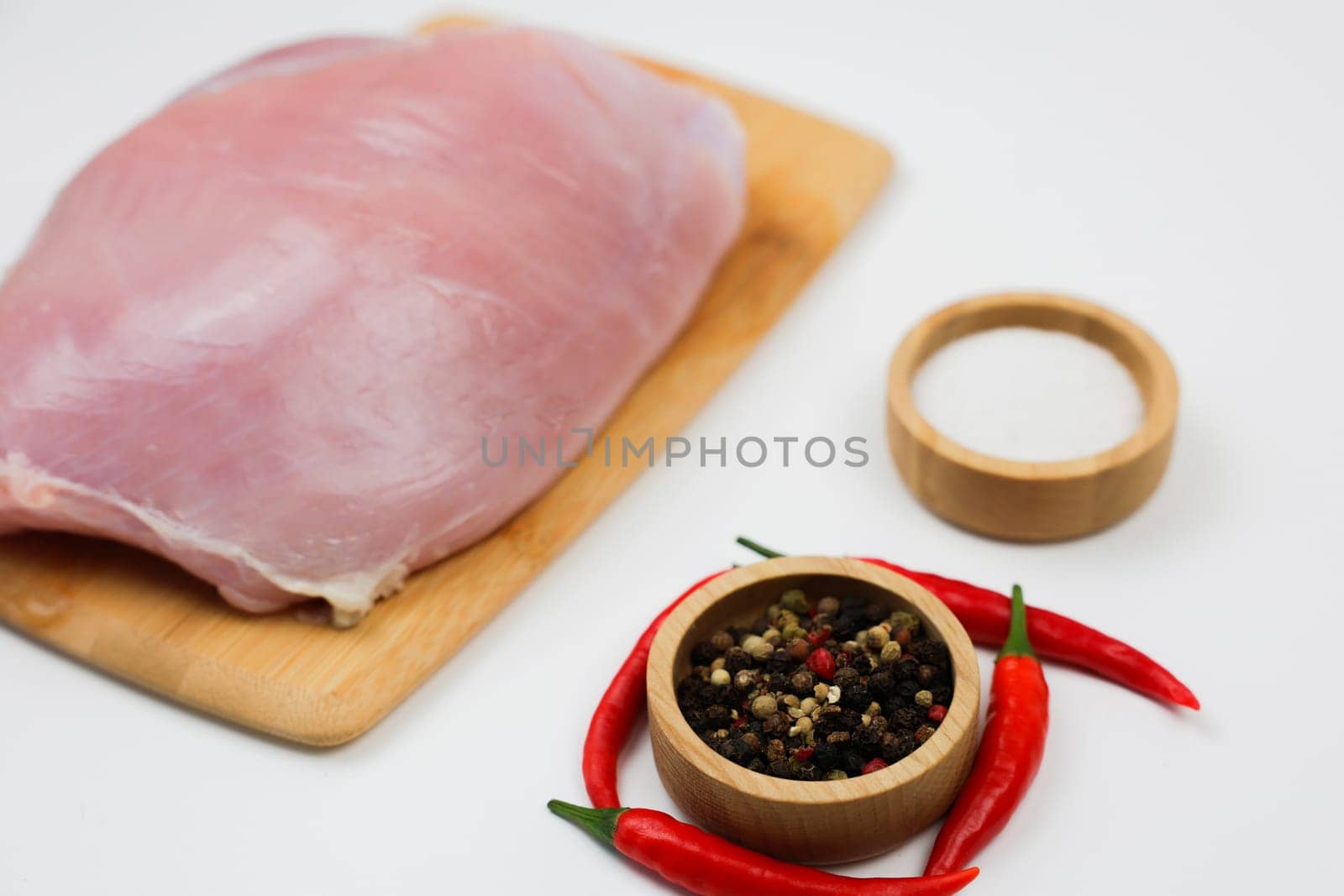 Close-up of raw turkey fillet lying on a wooden cutting board, next to spices and salt in wooden bowls and red hot chili peppers on a white background. The concept of dietary dishes, spicy food, healthy and natural products. High quality photo