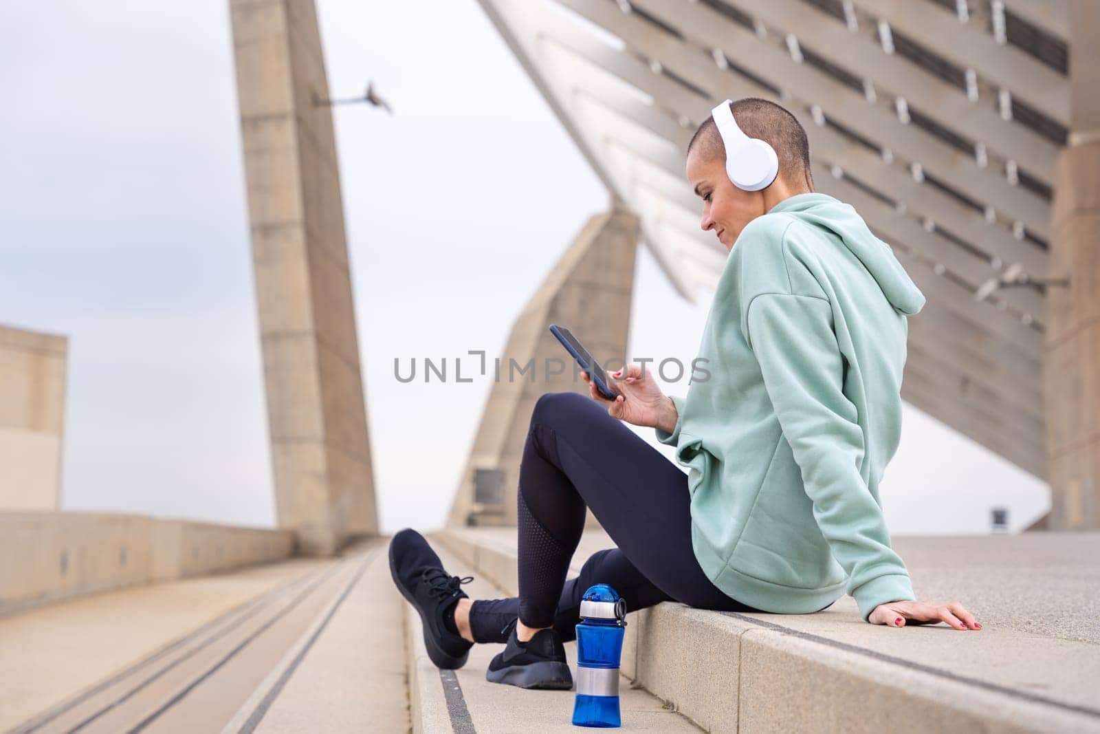 An athletic woman with headphones tired from exercising sits down to take a water break while using a cell phone app.