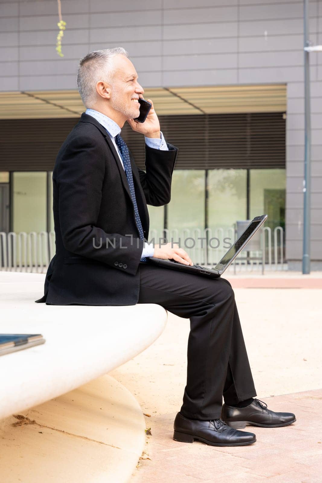 A middle-aged adult man in a suit has a business call while looking at his laptop.