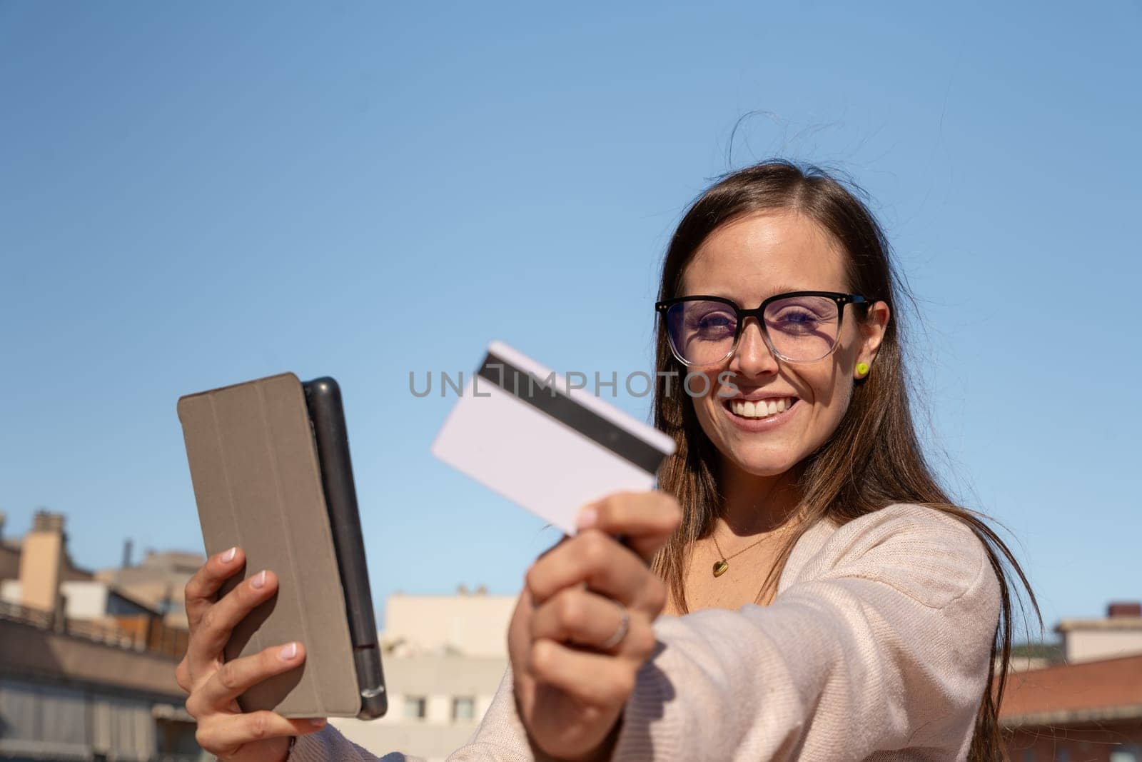 Happy young woman with glasses holding in each hand a credit card and digital tablet. Looking into the camera. Concept: payment in an online store
