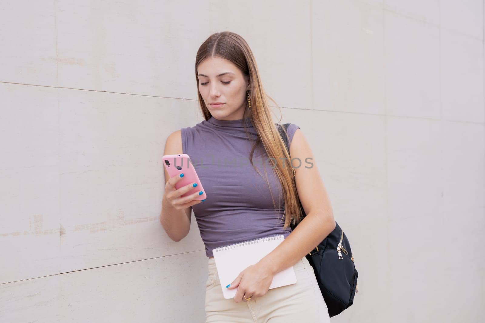 Front view of a laughing Caucasian teenage girl using a social media application with her mobile phone, leaning on the wall. University life concept