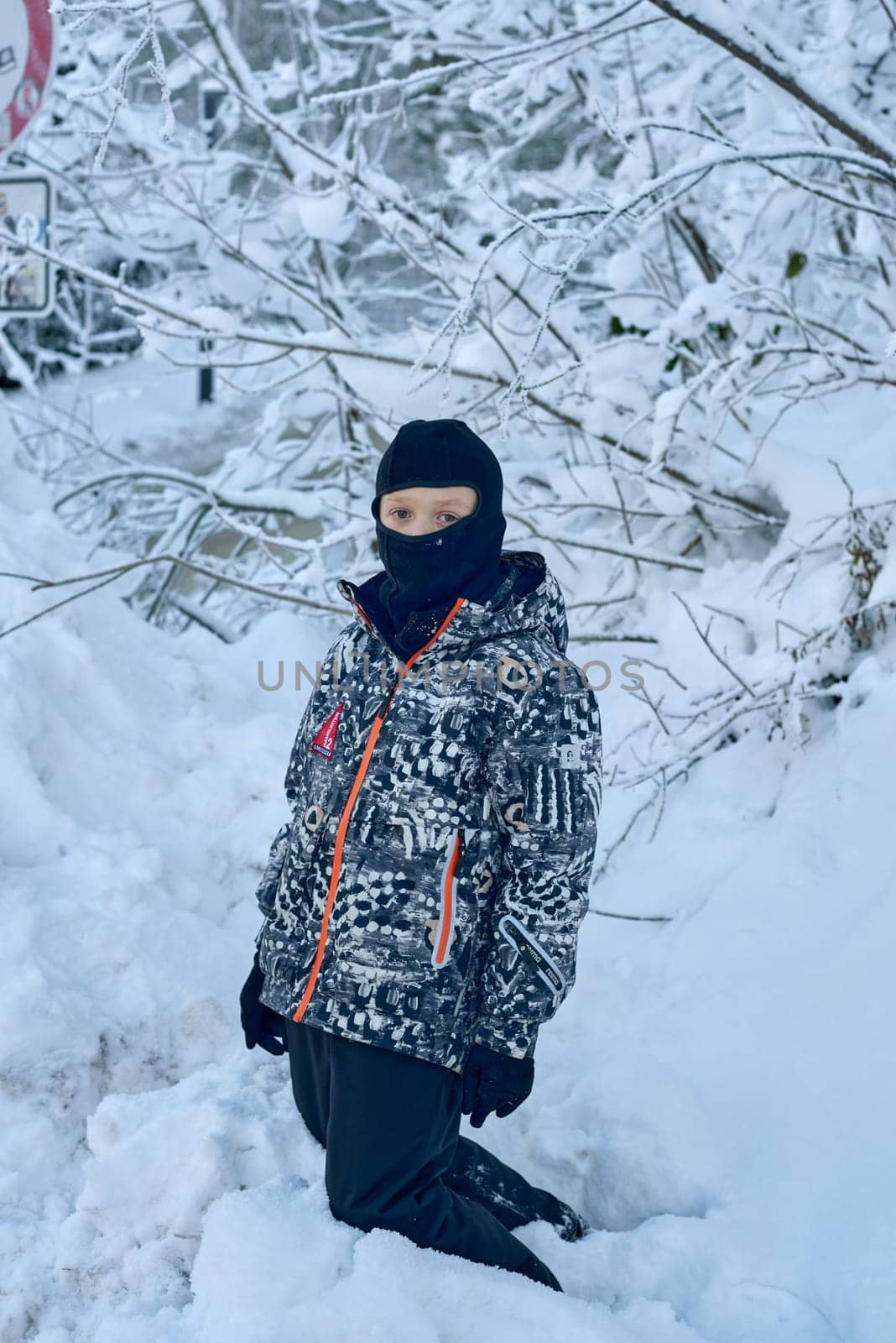 This high-resolution photograph captures a young boy, approximately 8 to 10 years old, kneeling in a serene, snow-covered forest. The boy is dressed warmly in a black hooded jacket featuring an abstract pattern of black, white, and gray swirls with bright orange accents. A black balaclava covers his head and face, leaving only his eyes and mouth visible, while black gloves complete his winter outfit. He kneels in a thick layer of undisturbed snow, surrounded by a dense forest of snow-laden branches that create intricate white patterns against the dark, leafless trees. A partially visible snow-covered signpost to the left hints at a winter sports area or restricted zone. The soft lighting and pristine snowy setting evoke a peaceful and reflective atmosphere, perfect for themes of winter exploration, childhood wonder, and outdoor adventure.