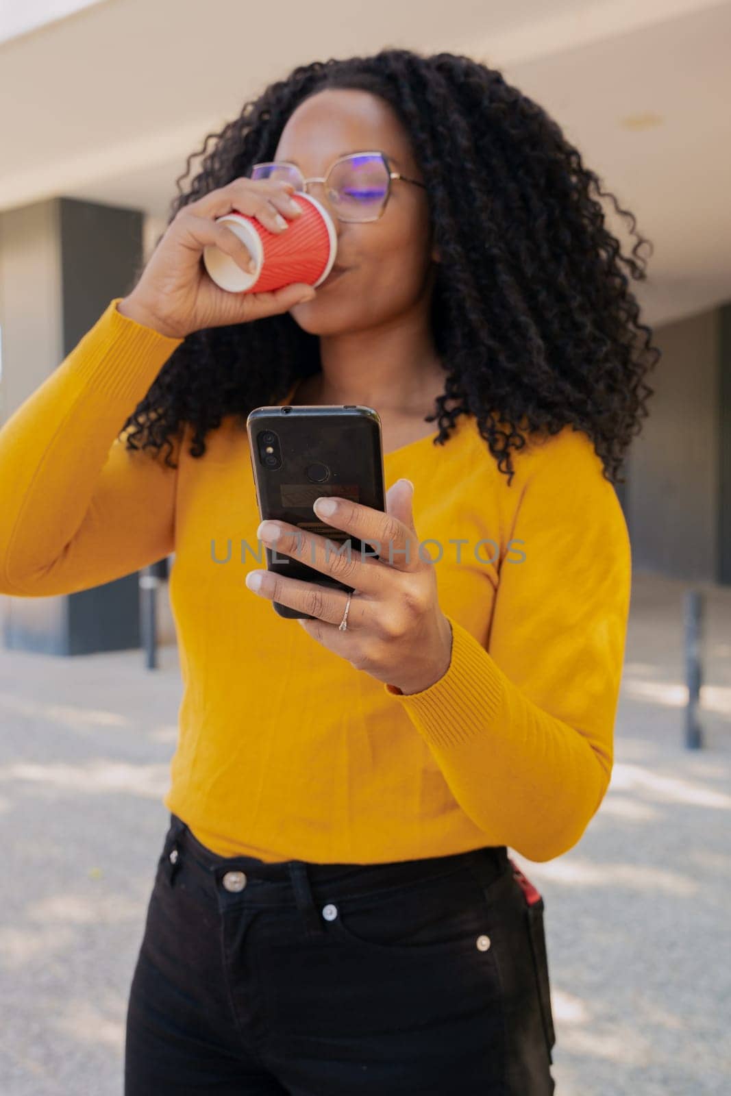 Vertical photo Young smiling black woman looking at social media on her mobile phone on campus while drinking coffee. concept: university life