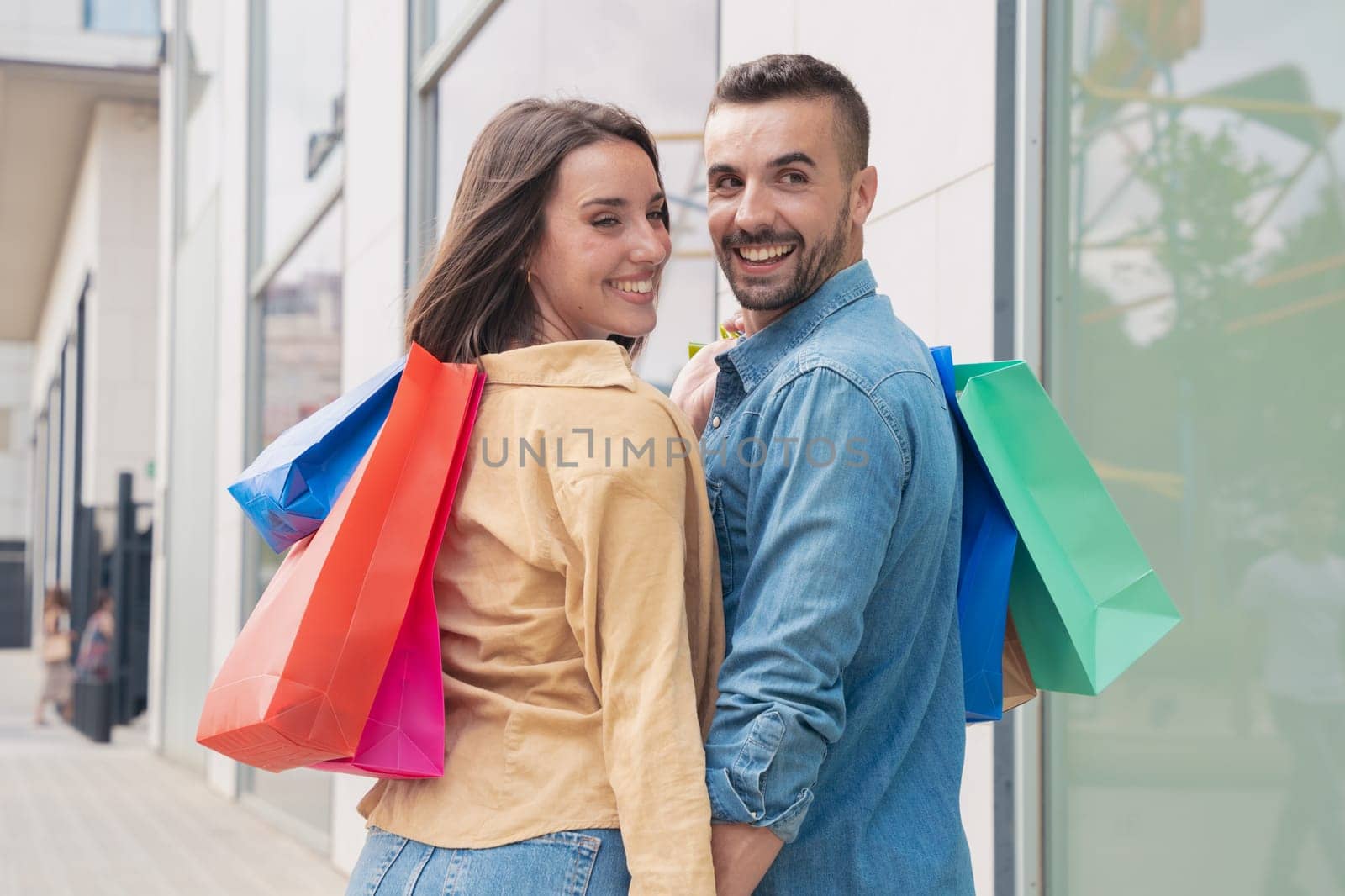 Young happy couple walking with shopping bags and turning back smiling to look and wave at the camera on an urban city street. Consumption concept.
