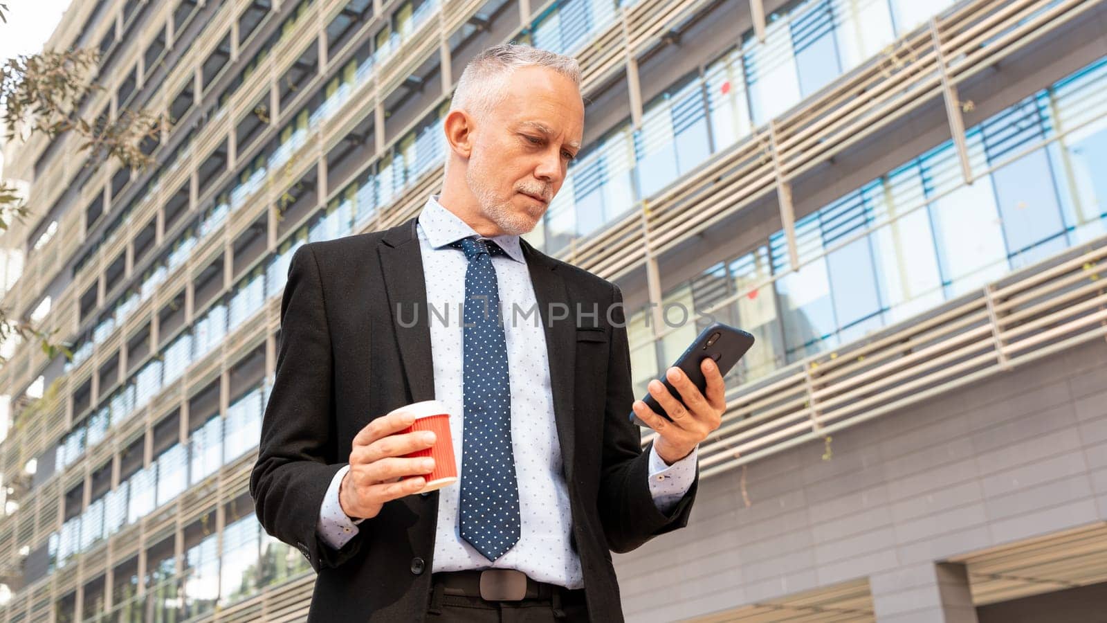 Satisfied with the results, the businessman walks down the street outside the office building, a mature boss holds a phone and coffee in his hands, writes messages and reads news online, using an app.