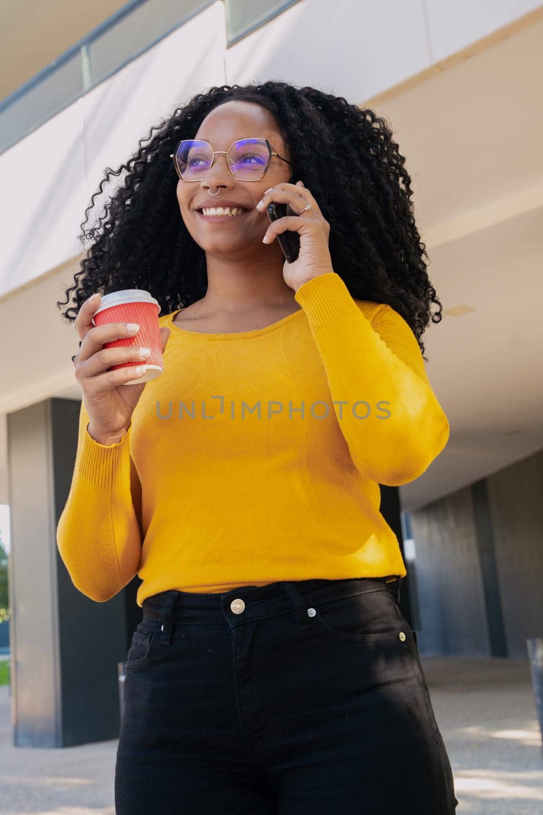 Vertical photo Young smiling black student talking on mobile phone while drinking coffee on university campus. Concept: university life