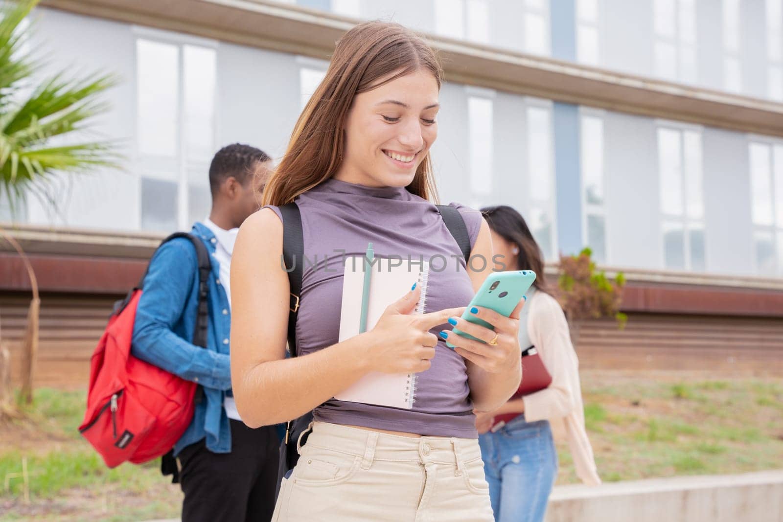 Young smiling Caucasian student with notebook in hand uses social networks with smartphone applications and wireless technology outdoors