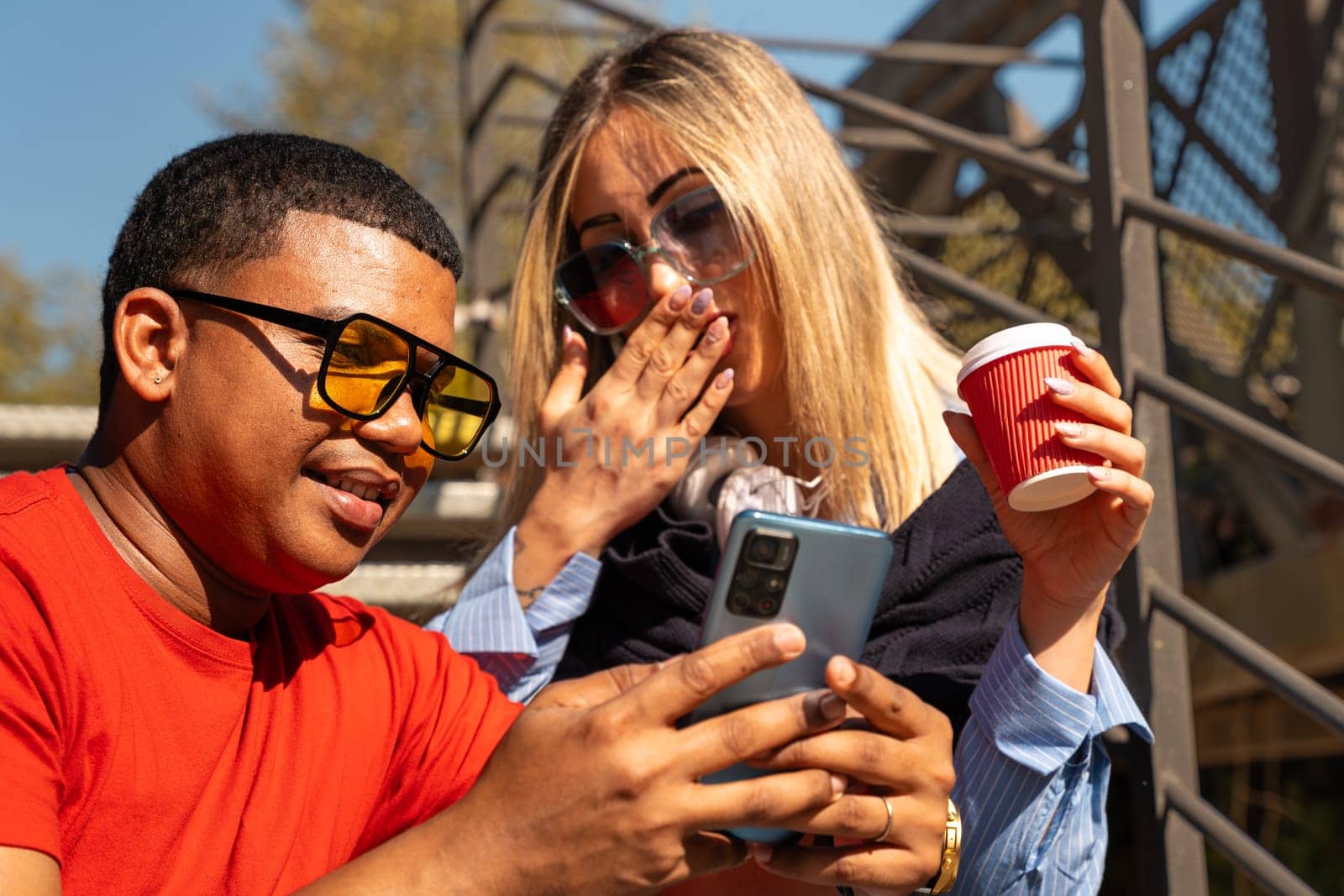 Happy diverse friends in the park sitting together on a bench surprised looking at social media on smartphones talking and smiling.