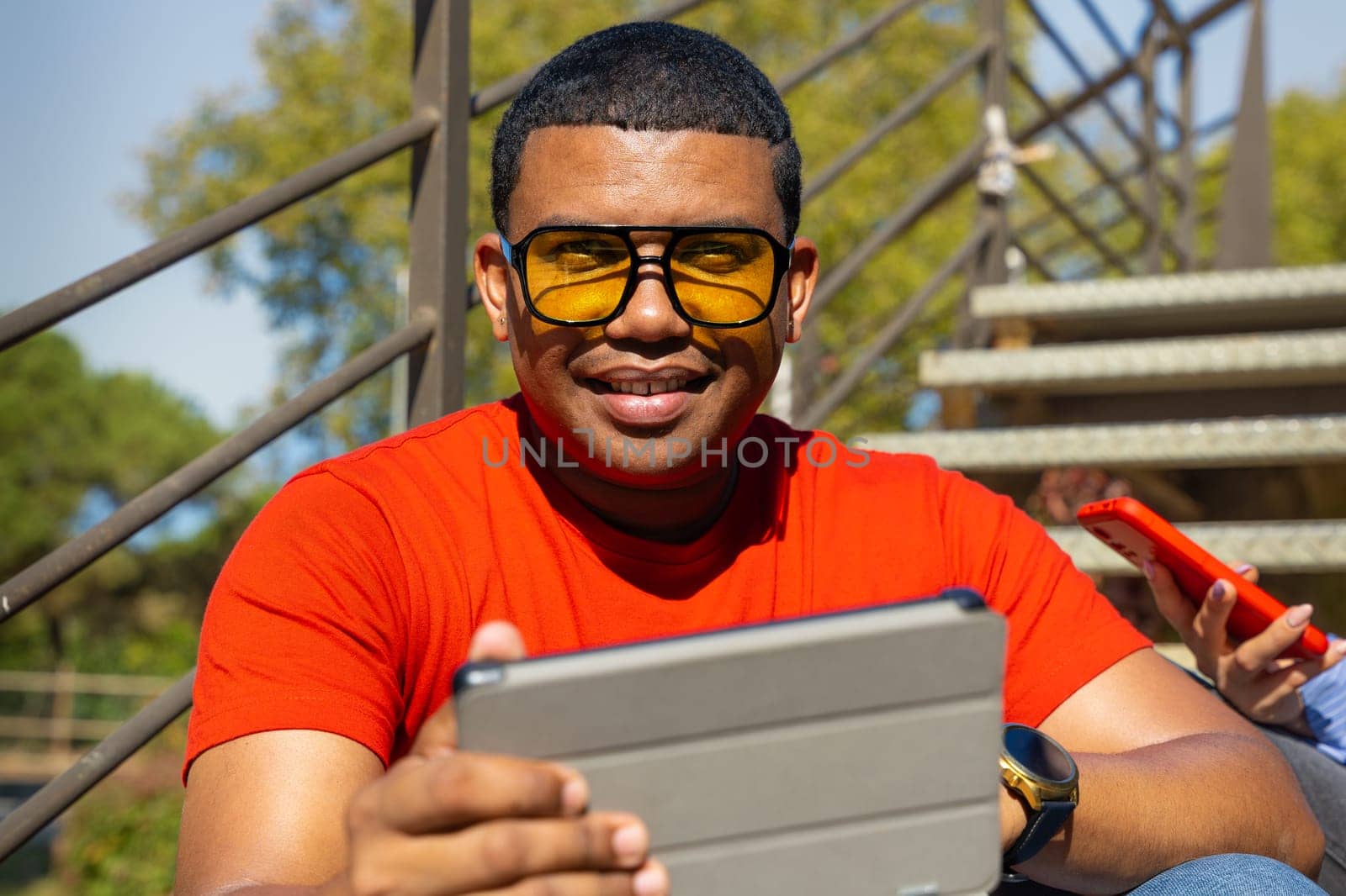 Happy latin man wearing sunglasses smiling while surfing the internet with digital tablet looking at camera outdoors.