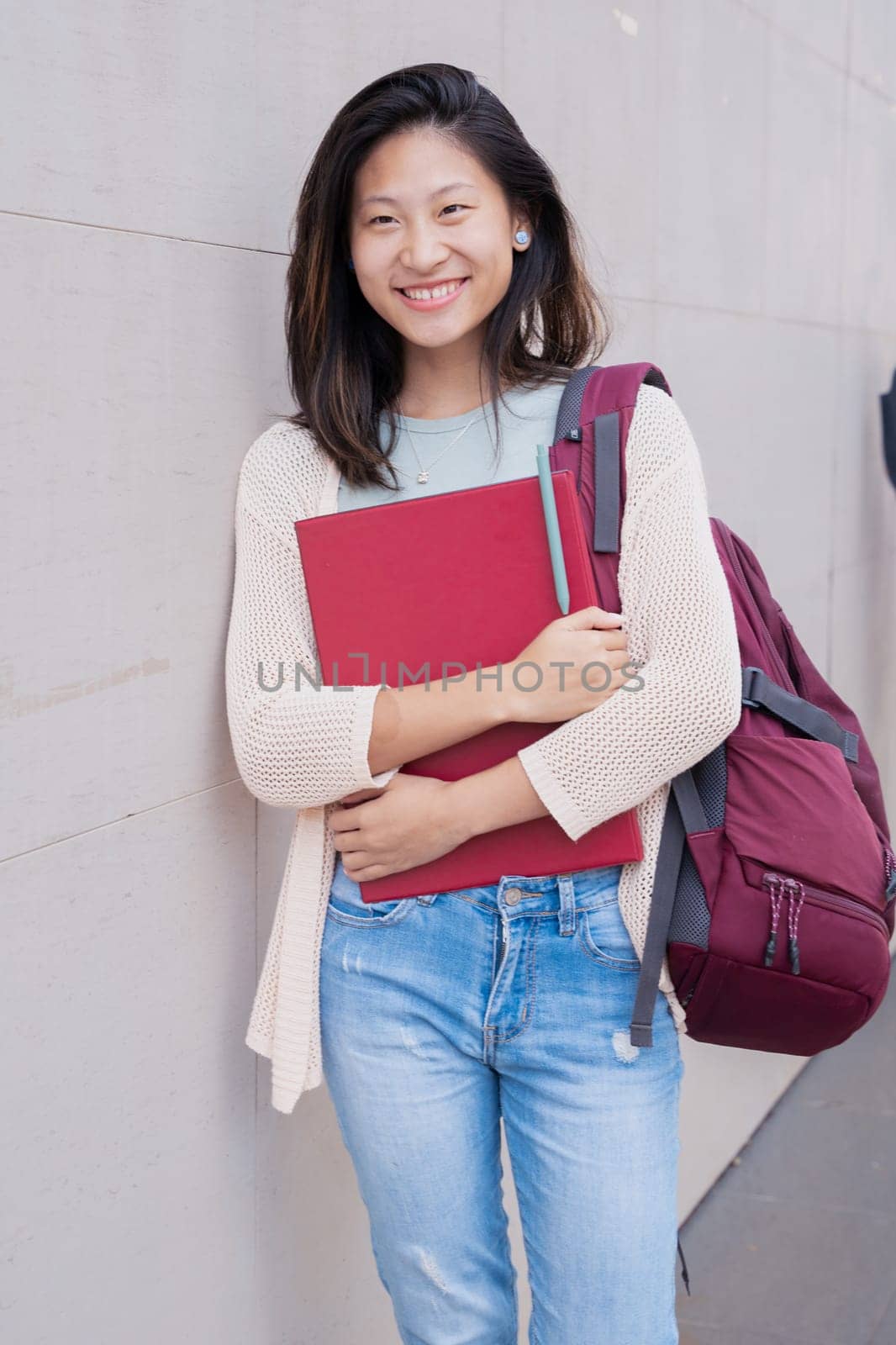 Front view of smiling Asian teenage girl with backpack leaning on the wall.College life concept