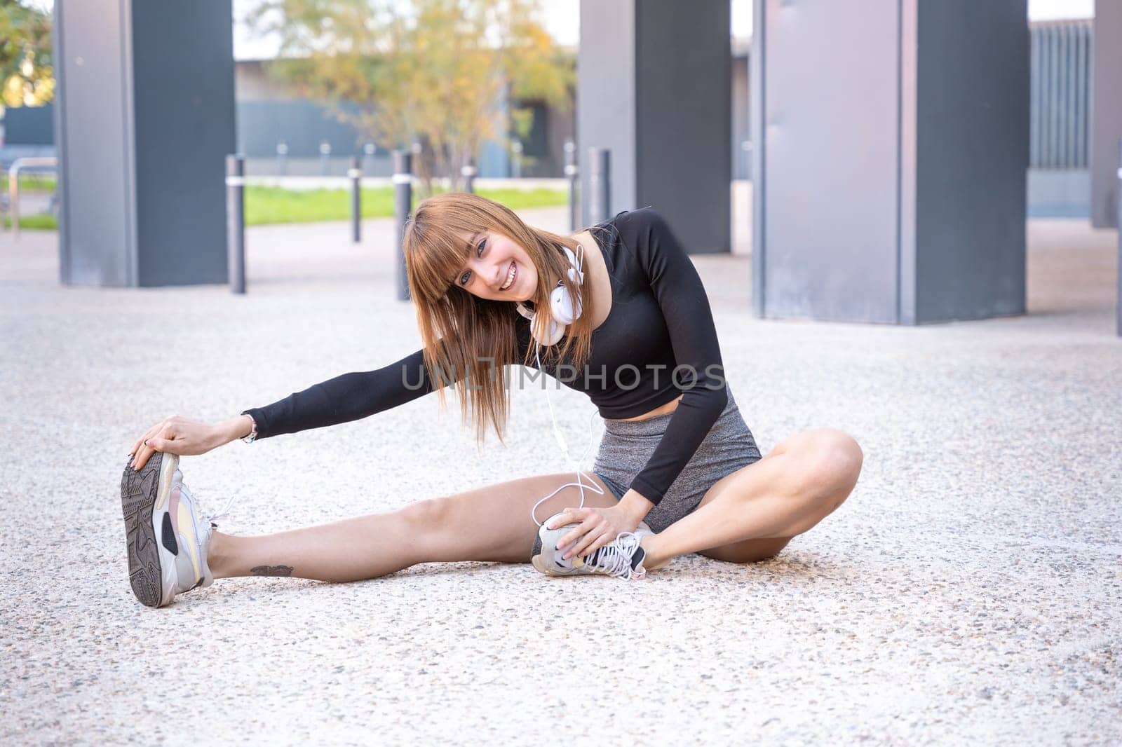 Sporty caucasian young woman wearing sports headphones doing stretches smiling looking at camera outdoors.
