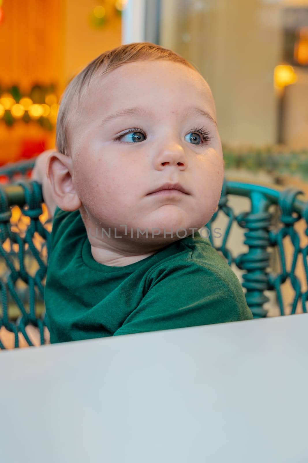Close-up portrait of a very beautiful boy with blonde hair and big green eyes sitting on the terrace in a chair.