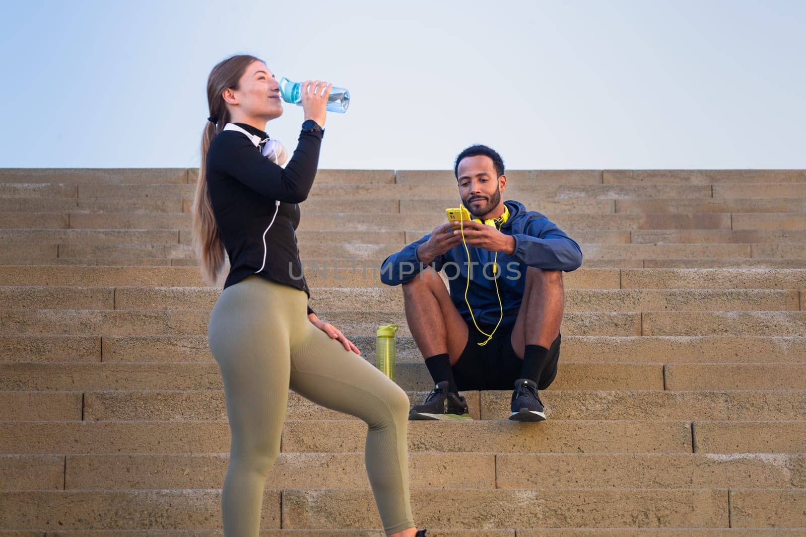 Smiling man using a social media app on a smartphone and young woman drinking water in sports bottle while exercising outdoors.