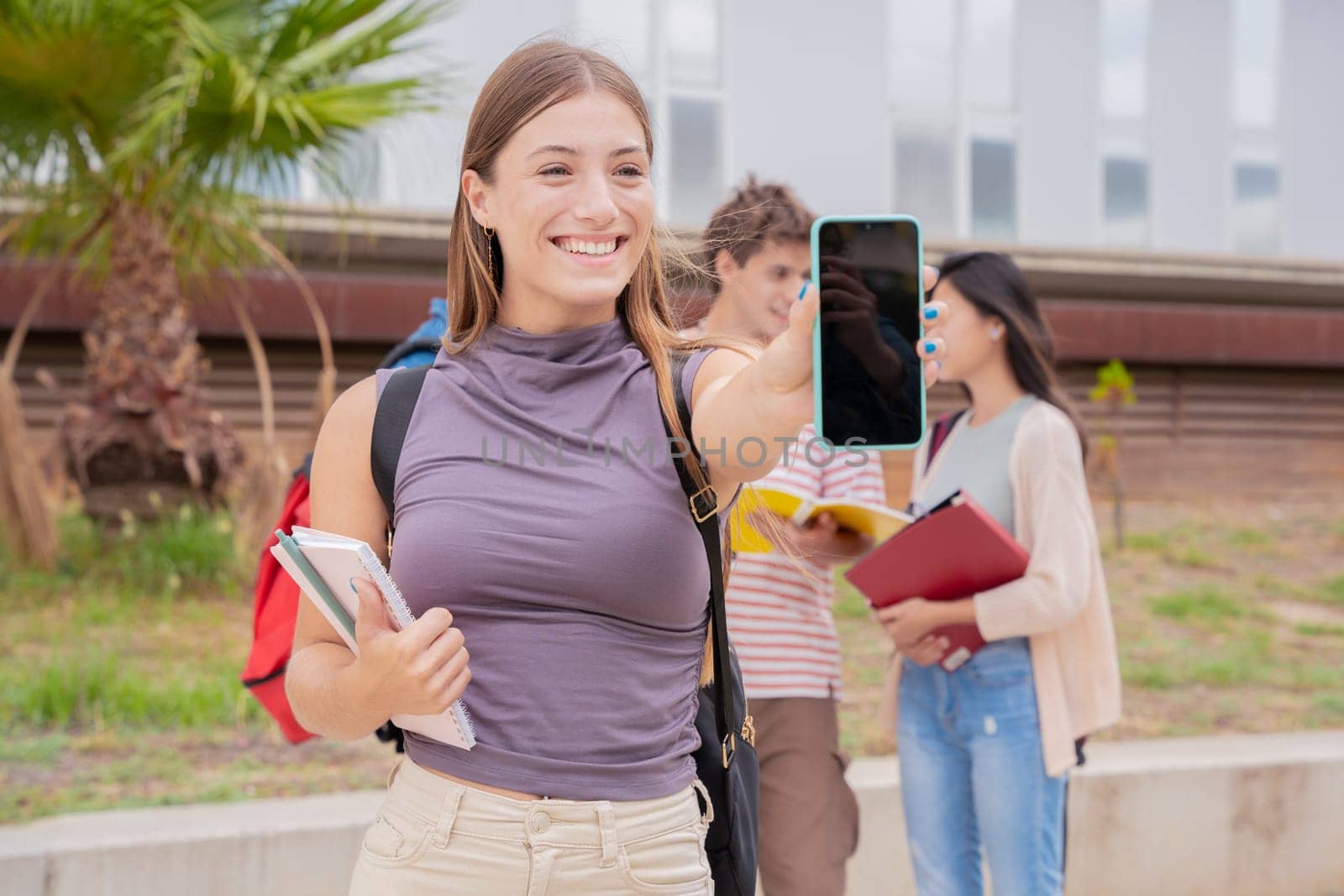 Young smiling student holding and showing an empty smartphone screen, behind her classmates talking and reviewing class notes.
