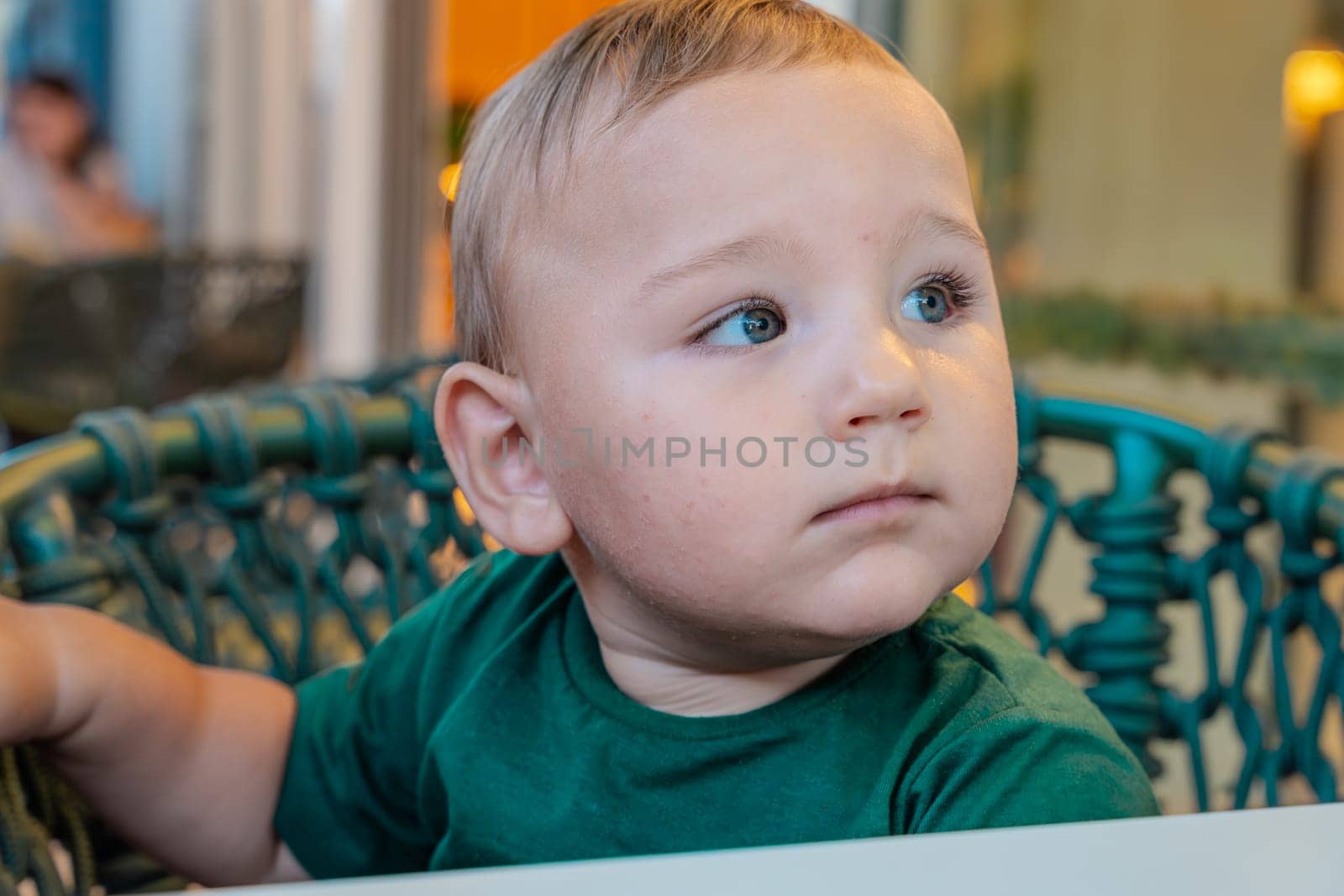 Close-up portrait of a very beautiful boy with blonde hair and big green eyes sitting on the terrace in a chair.