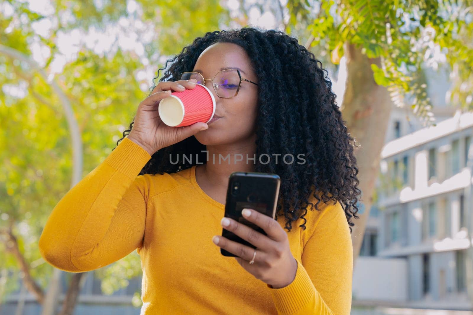 Young smiling black woman student looking at social media on her mobile phone on campus while drinking coffee. concept:university life
