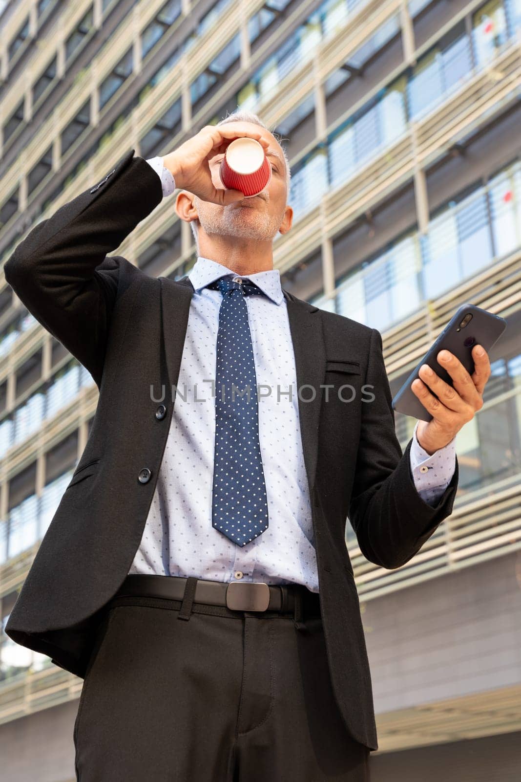 Satisfied with the results, the businessman walks down the street outside the office building, a mature boss holds a phone and coffee in his hands, writes messages and reads news online, using an app.