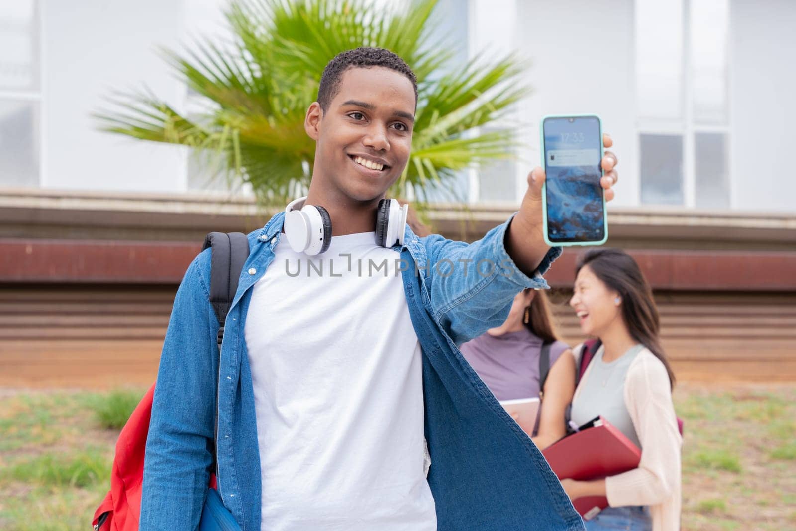 Young smiling black student holding and showing an empty smartphone screen, behind her classmates talking and reviewing class notes.