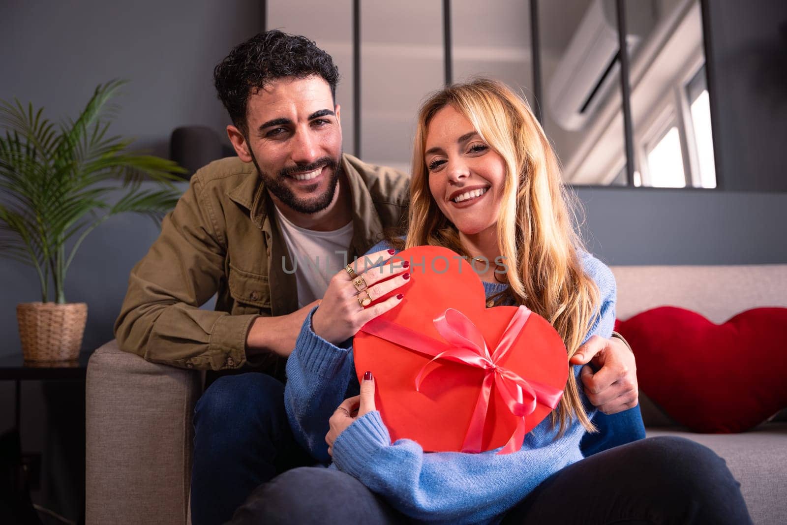 Beautiful young couple at home enjoying time together while celebrating Valentine's Day with a heart-shaped gift box in hand. Looking at the camera.