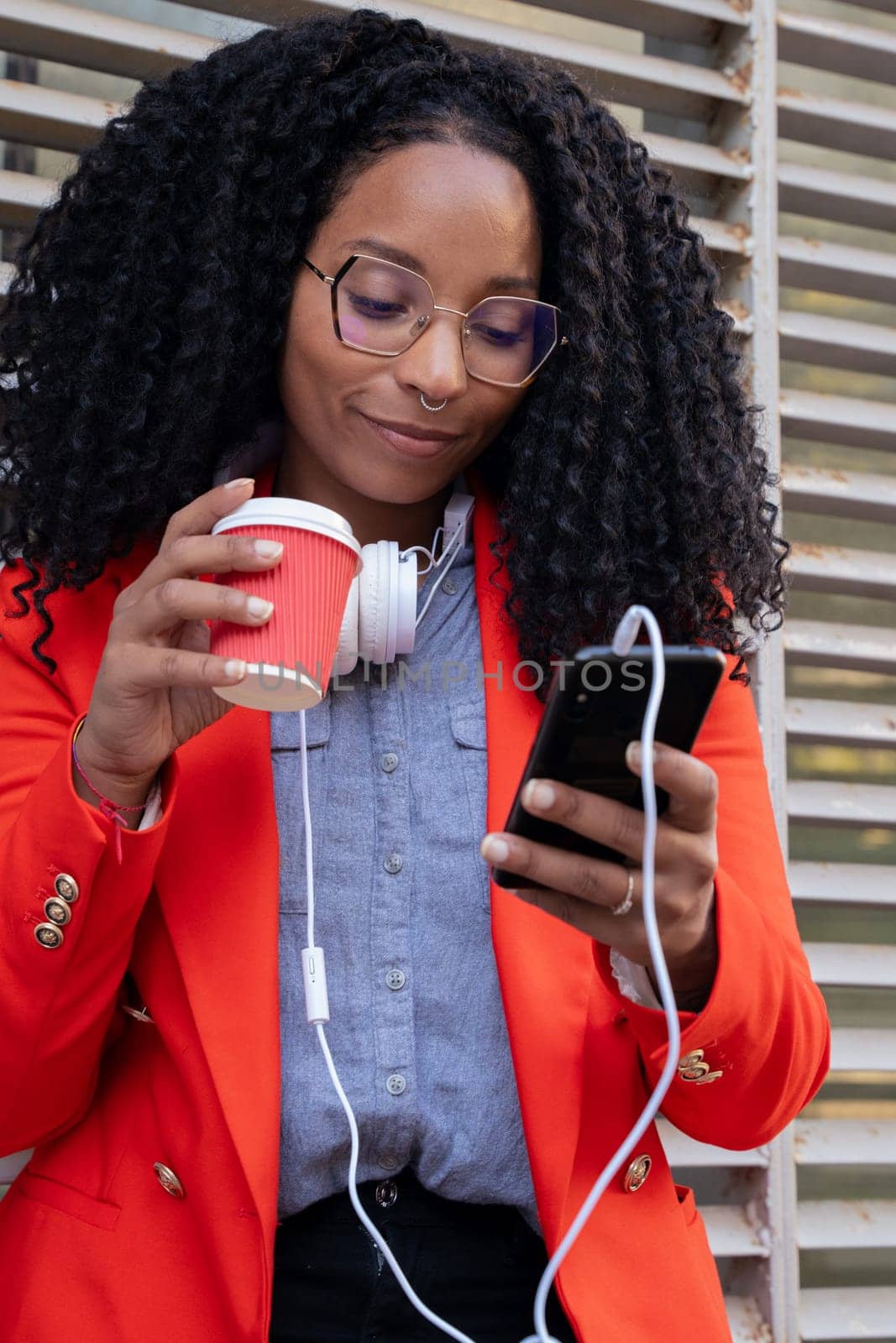 Black business woman with coffee in hand and headphones hanging around her neck, watching news with her mobile phone.