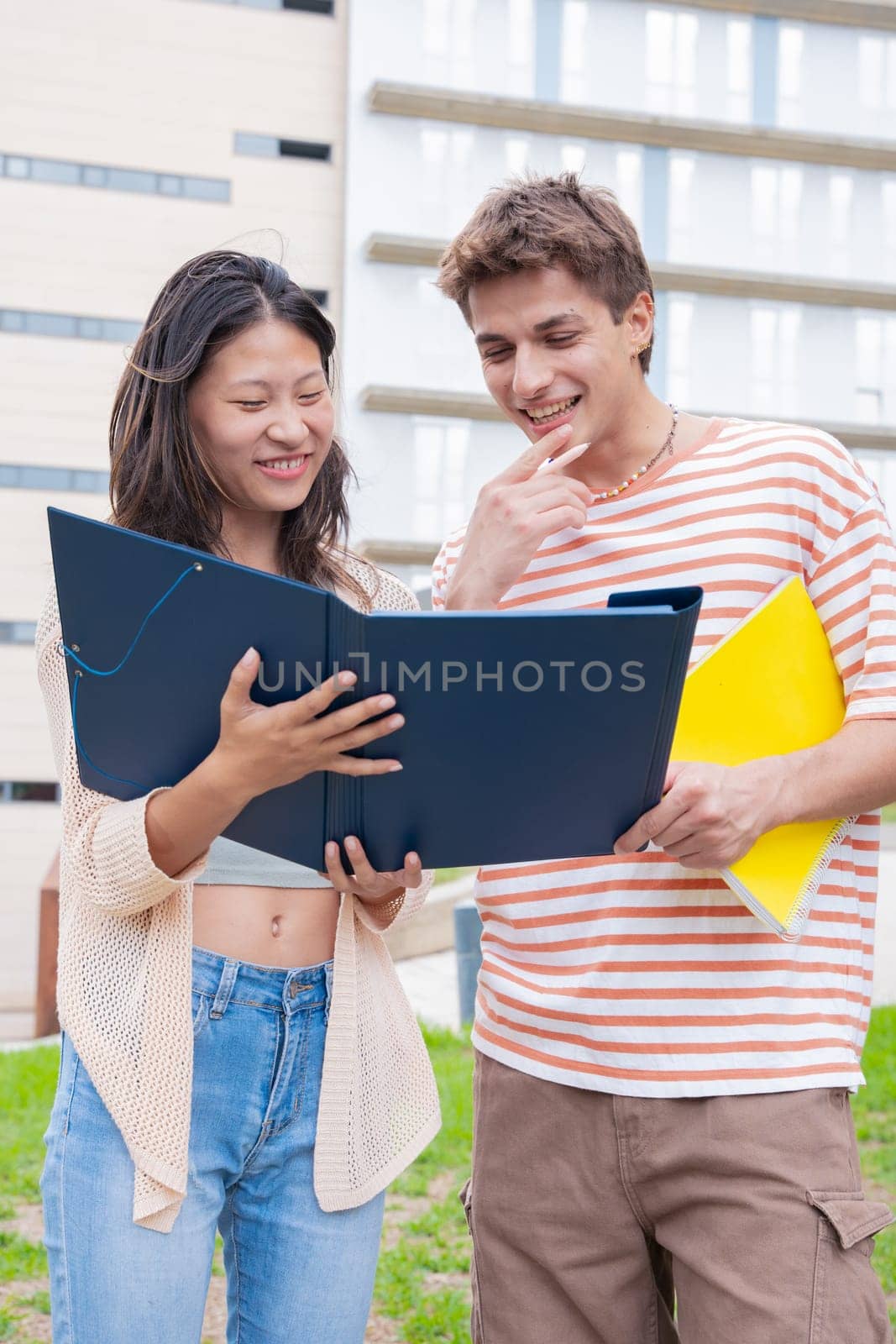 Happy university student friends talking and laughing while reviewing syllabus notes on campus. Diverse Asian and European youth
