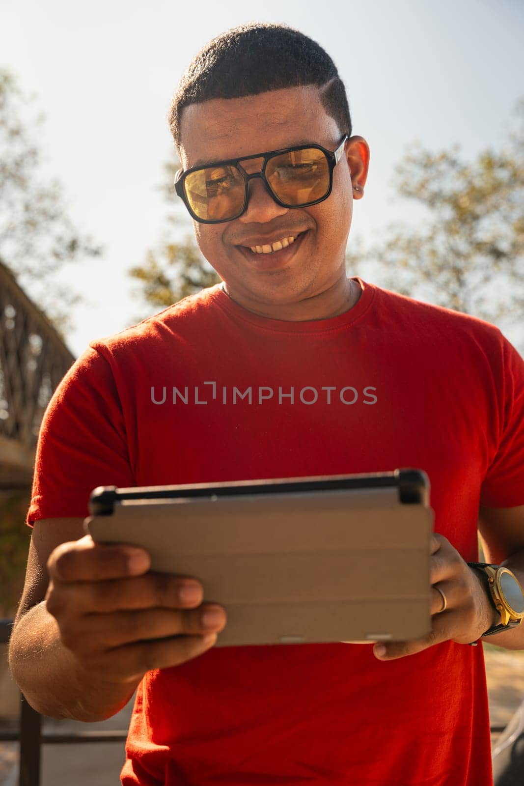 Happy latin man wearing sunglasses smiling while surfing the internet with digital tablet looking at camera outdoors.
