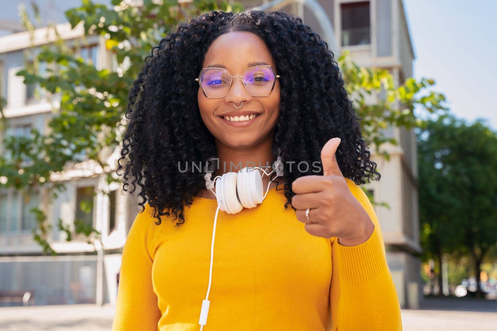 Close-up of happy curly haired young woman with glasses pointing thumb up. Facial expression of joy.