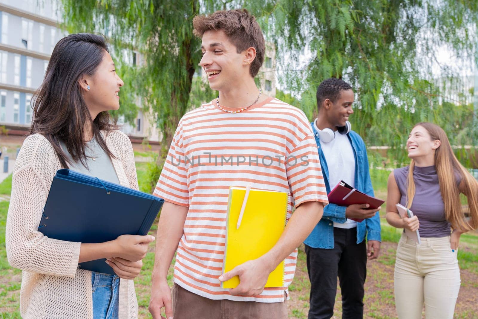 Happy university student friends talking and laughing while reviewing syllabus notes on campus. Diverse Asian and European youth
