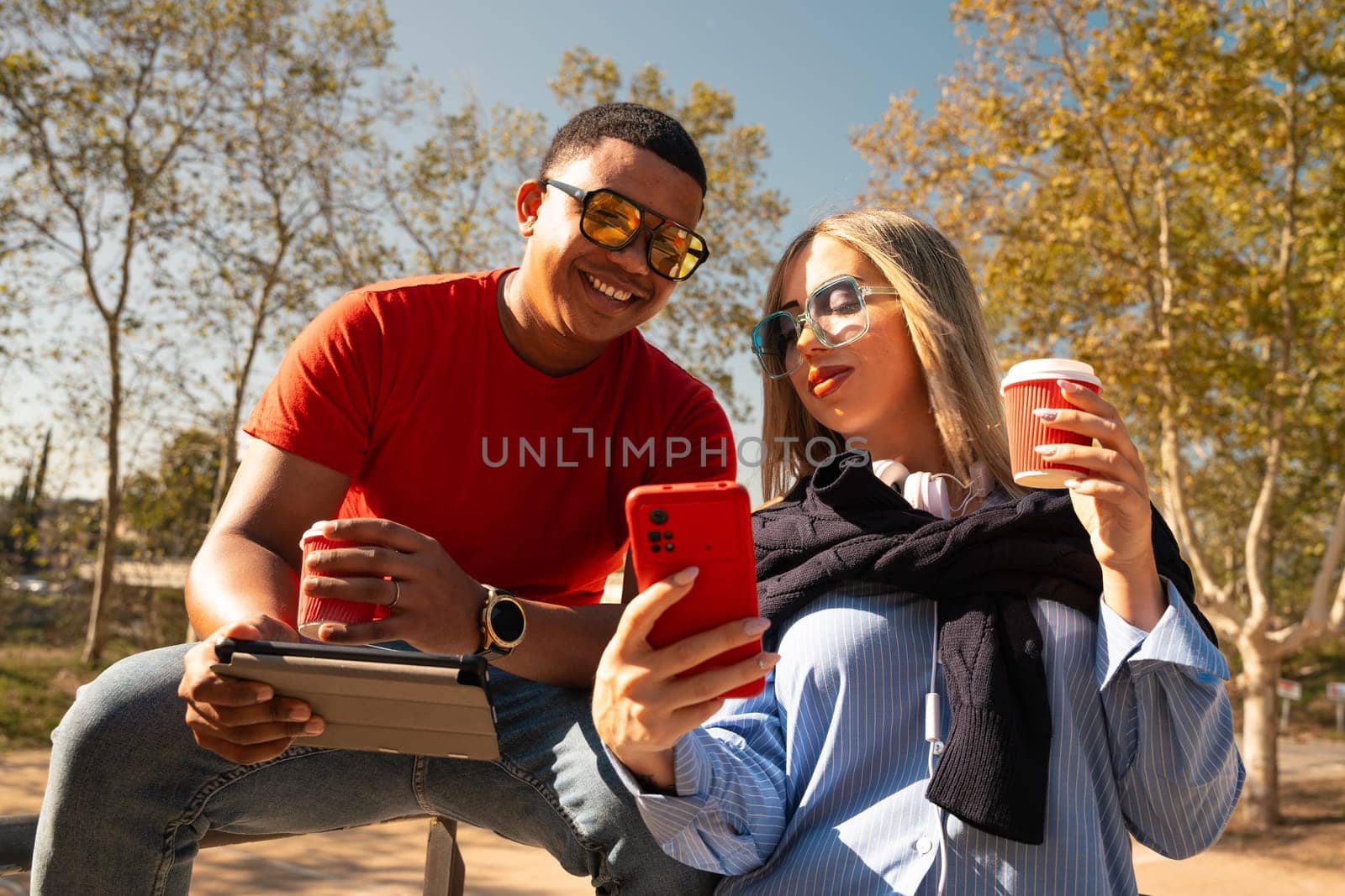 Happy diverse friends in the park sitting on a bench together looking at social media on smartphones talking and smiling.