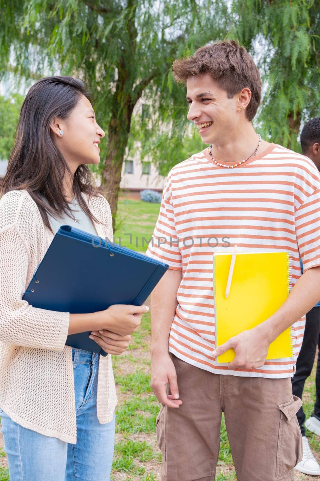 Happy university student friends talking and laughing while reviewing syllabus notes on campus. Diverse Asian and European youth