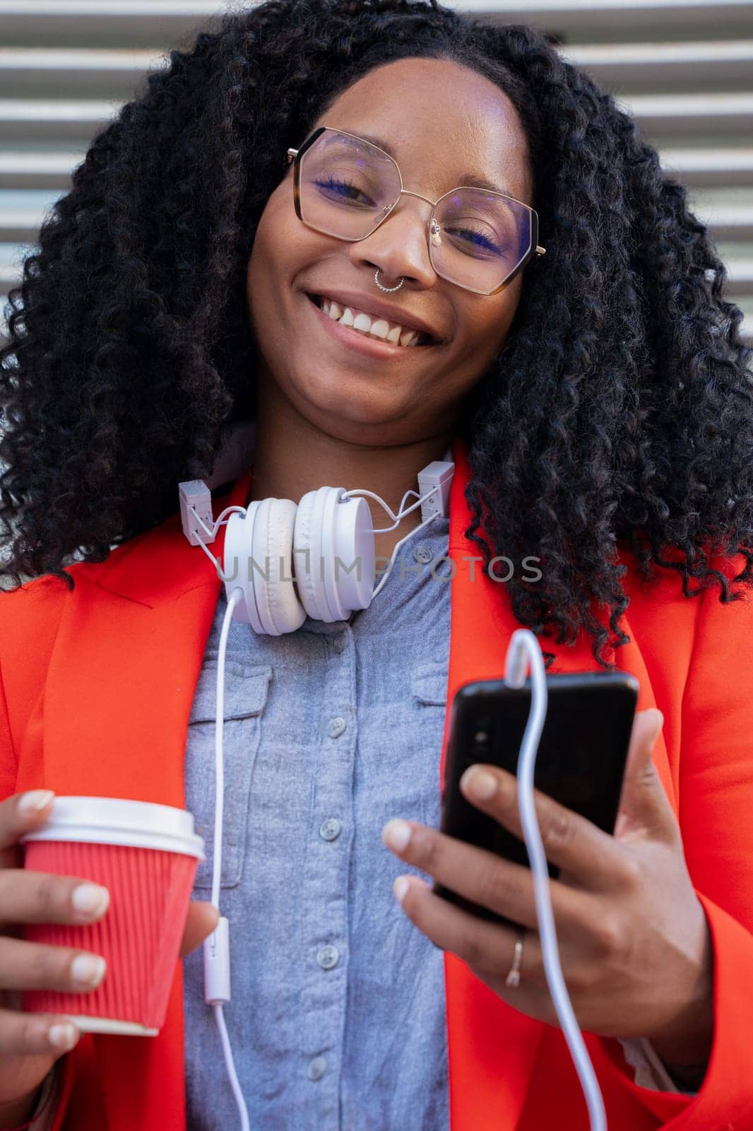 Portrait of a young smiling black business woman with mobile phone and coffee in her hands and headphones hanging around her neck.