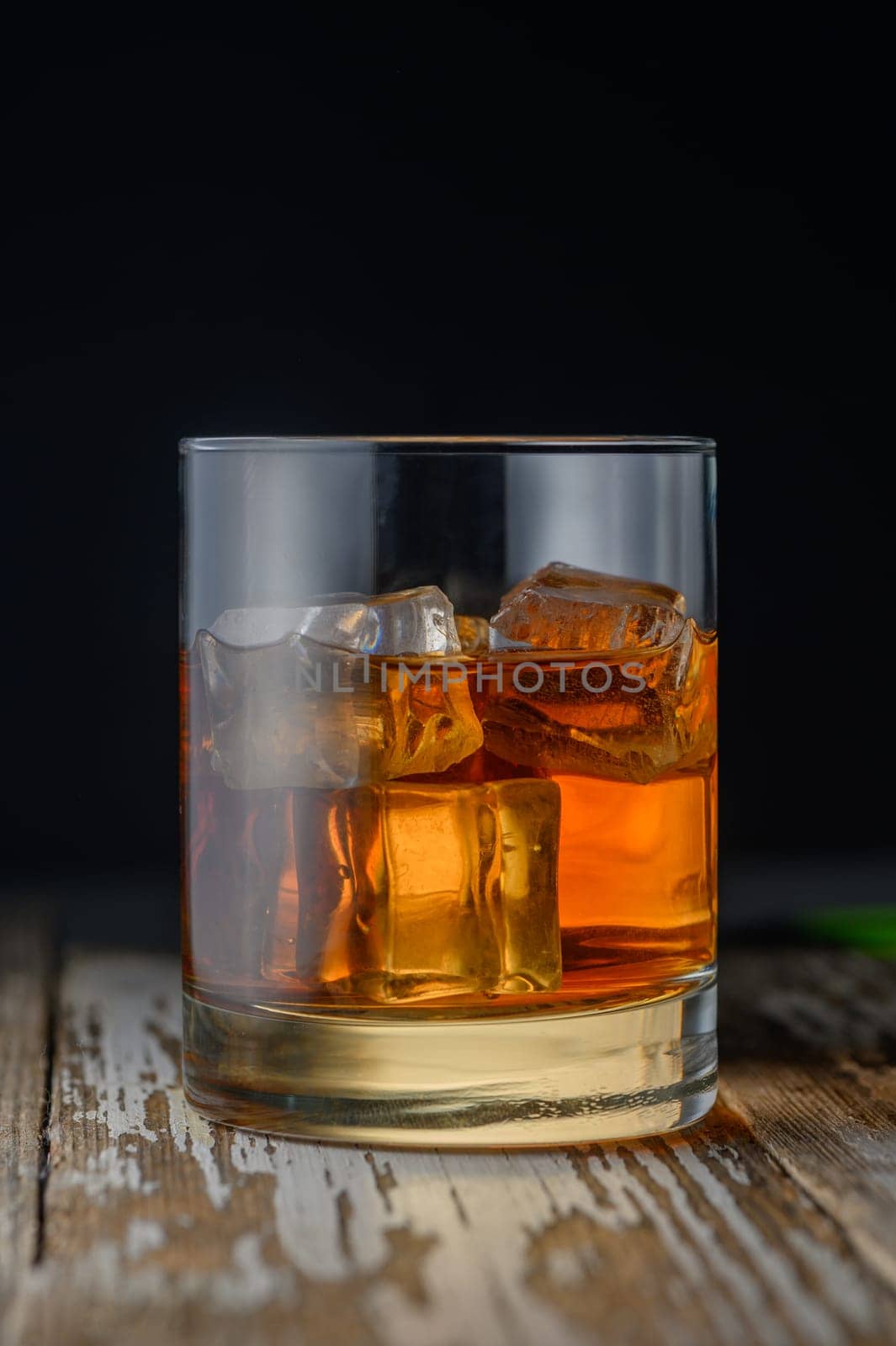 A glass filled with a golden drink and ice sits on a rustic wooden table, glistening under soft light.