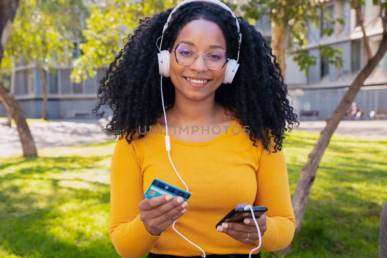 happy black woman wearing headphones pays with credit card on smartphone. Looking to the camera. Concept: online store payment