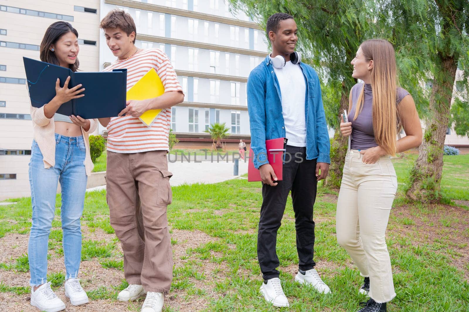 Happy university student friends talking and laughing while reviewing syllabus notes on campus. Diverse Asian and European youth