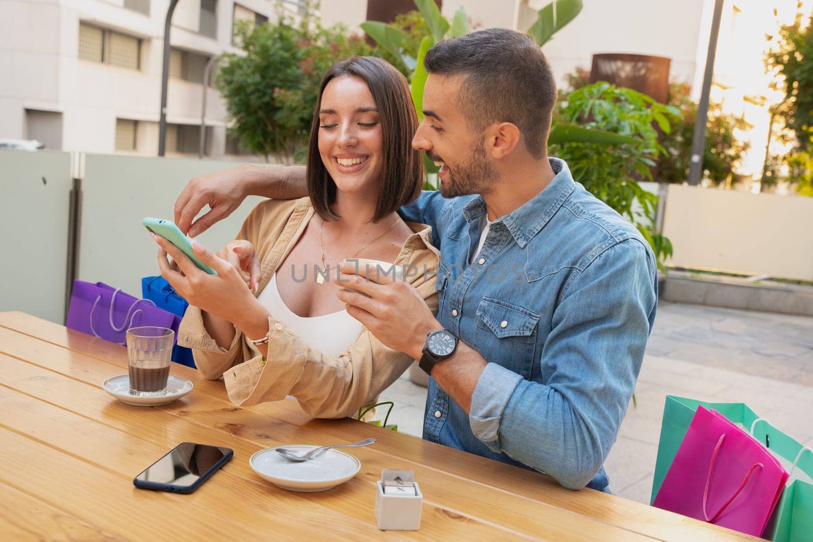 Sale, shopping, consumerism, leisure and people concept - happy couple looking at app on mobile phone shopping bags drinking coffee at shopping center