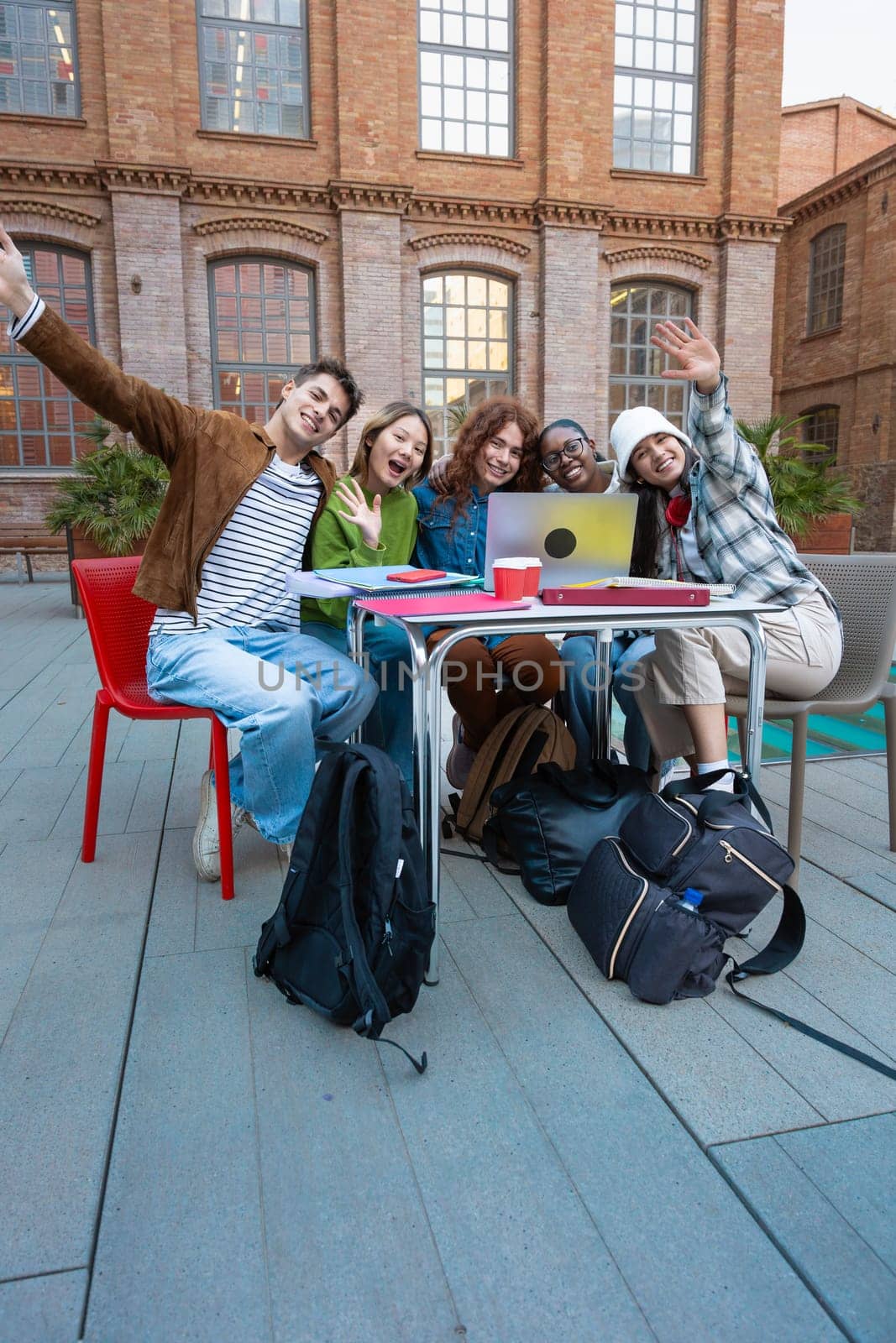 A multiethnic group of five students with a computer and notes sit near campus to review the exam outdoors. Looking at the camera
