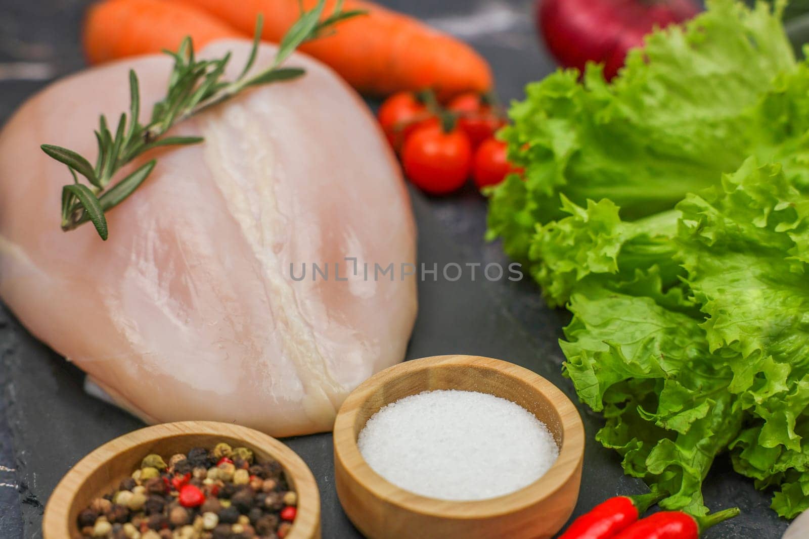 Close-up of raw chicken fillet with a sprig of rosemary lying surrounded by vegetables, herbs, spices on a dark background. The concept of diet, detox, proper and healthy nutrition, recipes. High quality photo