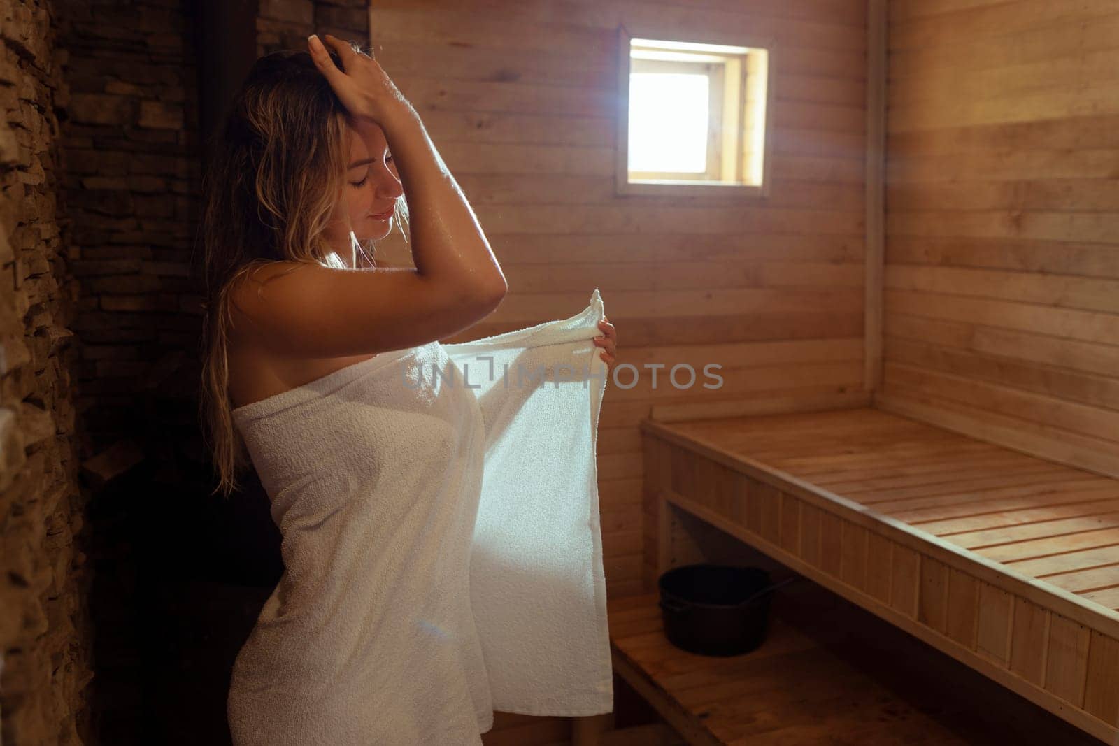 A young woman relaxes and sweats in a hot sauna wrapped in a towel. girl in the sauna. Finnish sauna interior, classic wooden sauna with hot steam. Russian bath