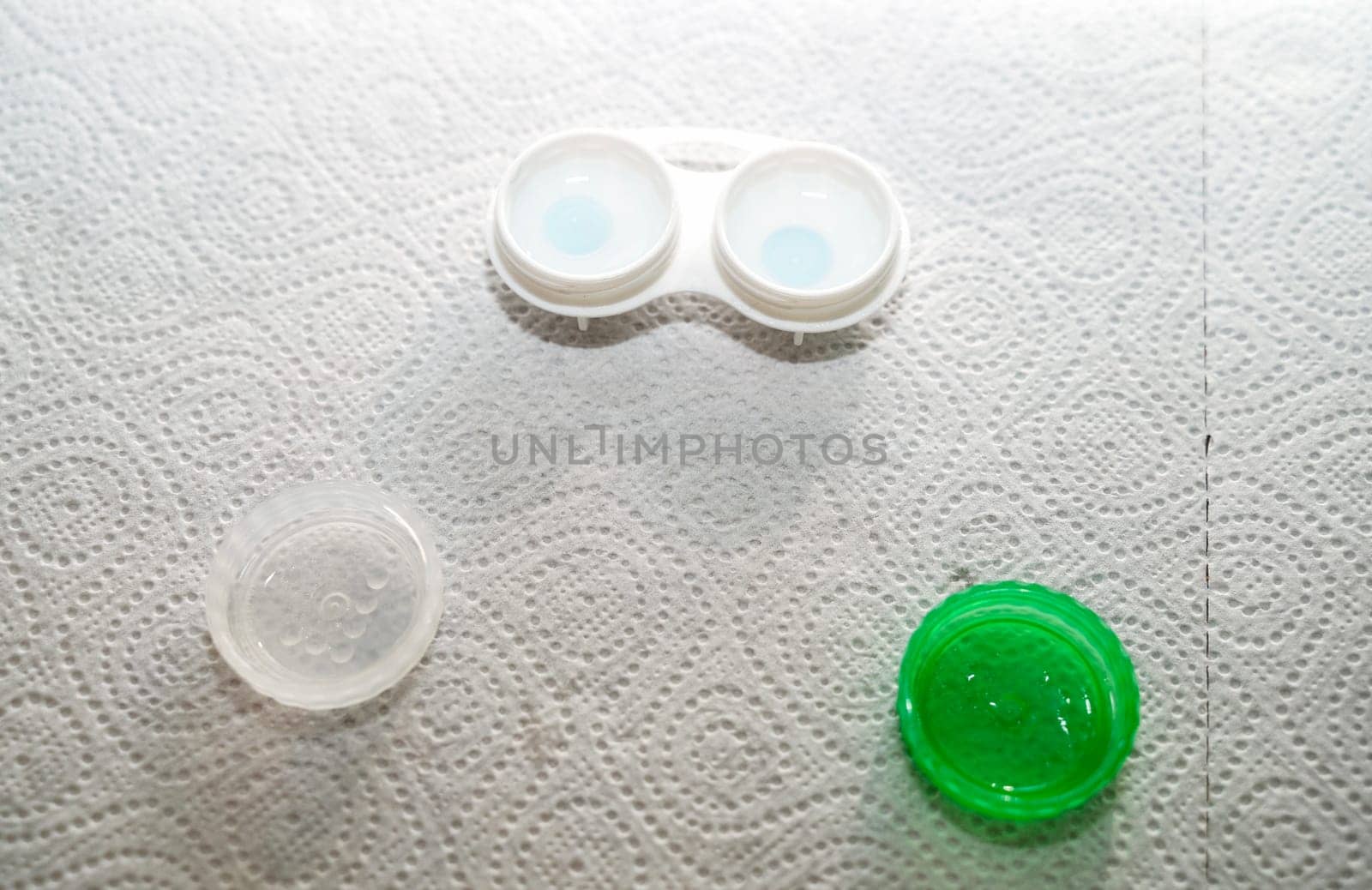 Container for storing lenses and contact lenses. The container is white with green, there are lenses in it. The table is covered with a paper towel for sterility at home. The container is open, you can see the lenses in it.