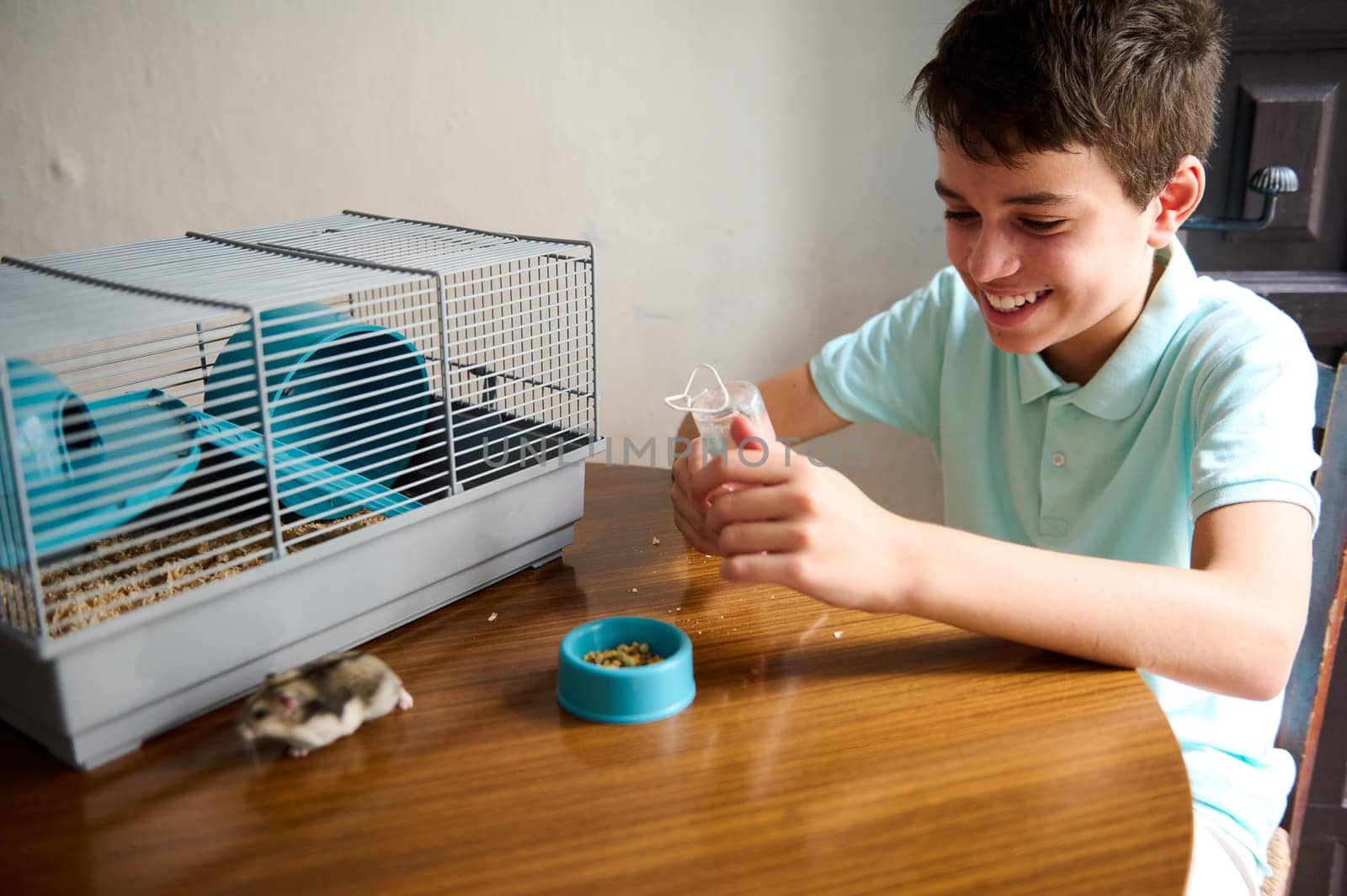 A cheerful boy sits at a table with his hamster and its cage, enjoying quality time together. The scene captures joy and care between a pet owner and his adorable, furry friend.